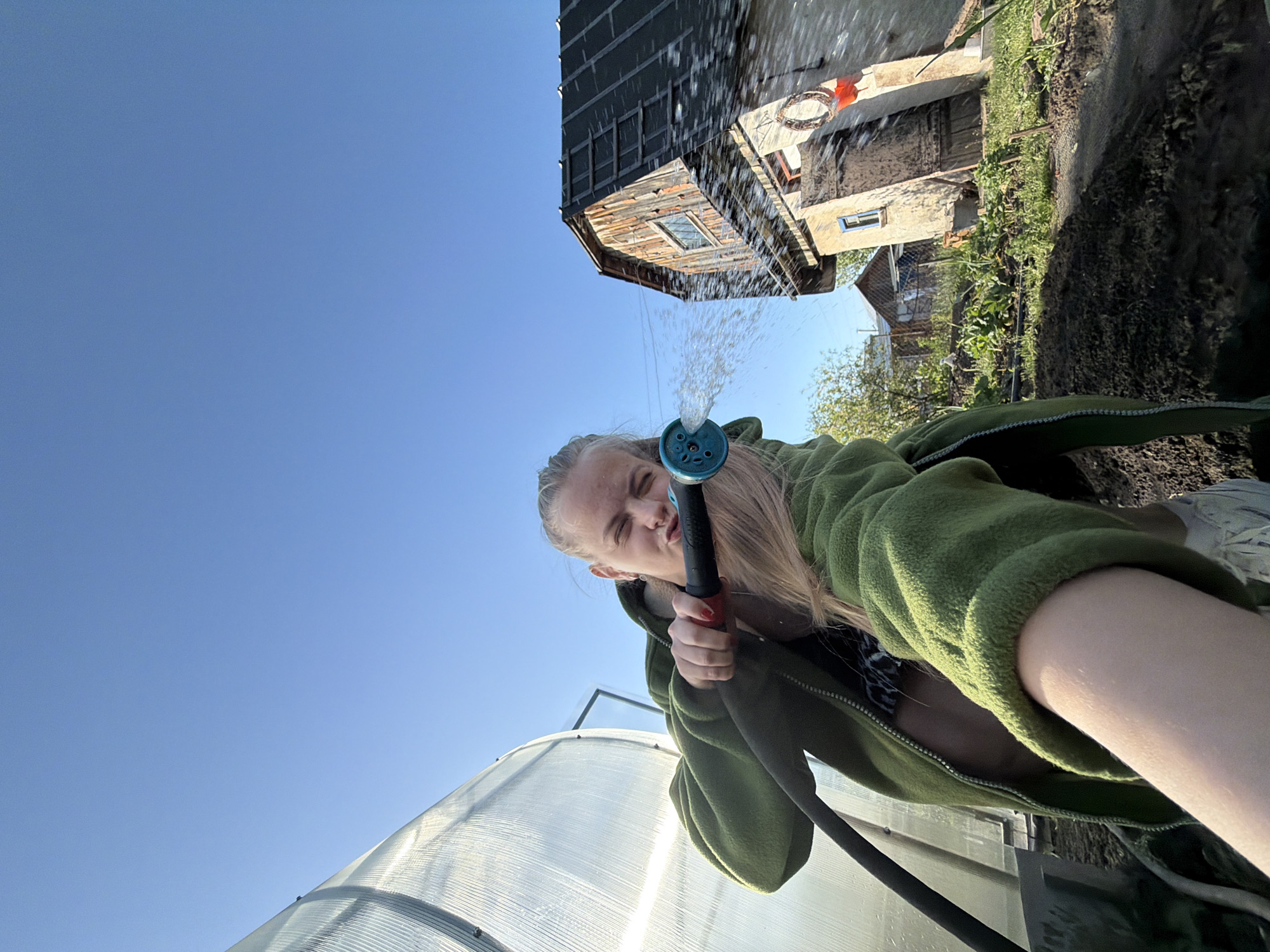 A low-angle, tilted, full shot depicts a young woman holding a blue and black garden hose, water spraying from its nozzle. She wears a green fleece jacket, partially unzipped, over a black and white patterned top. The sky is clear blue. A translucent greenhouse is visible in the lower left corner. In the background, there is a two-story building with gray siding and a window, as well as other structures and greenery. It is likely that the city of Chaikovsky is near the location because the model has geotagged herself there. The scene appears to be outdoors, possibly in a garden or yard.