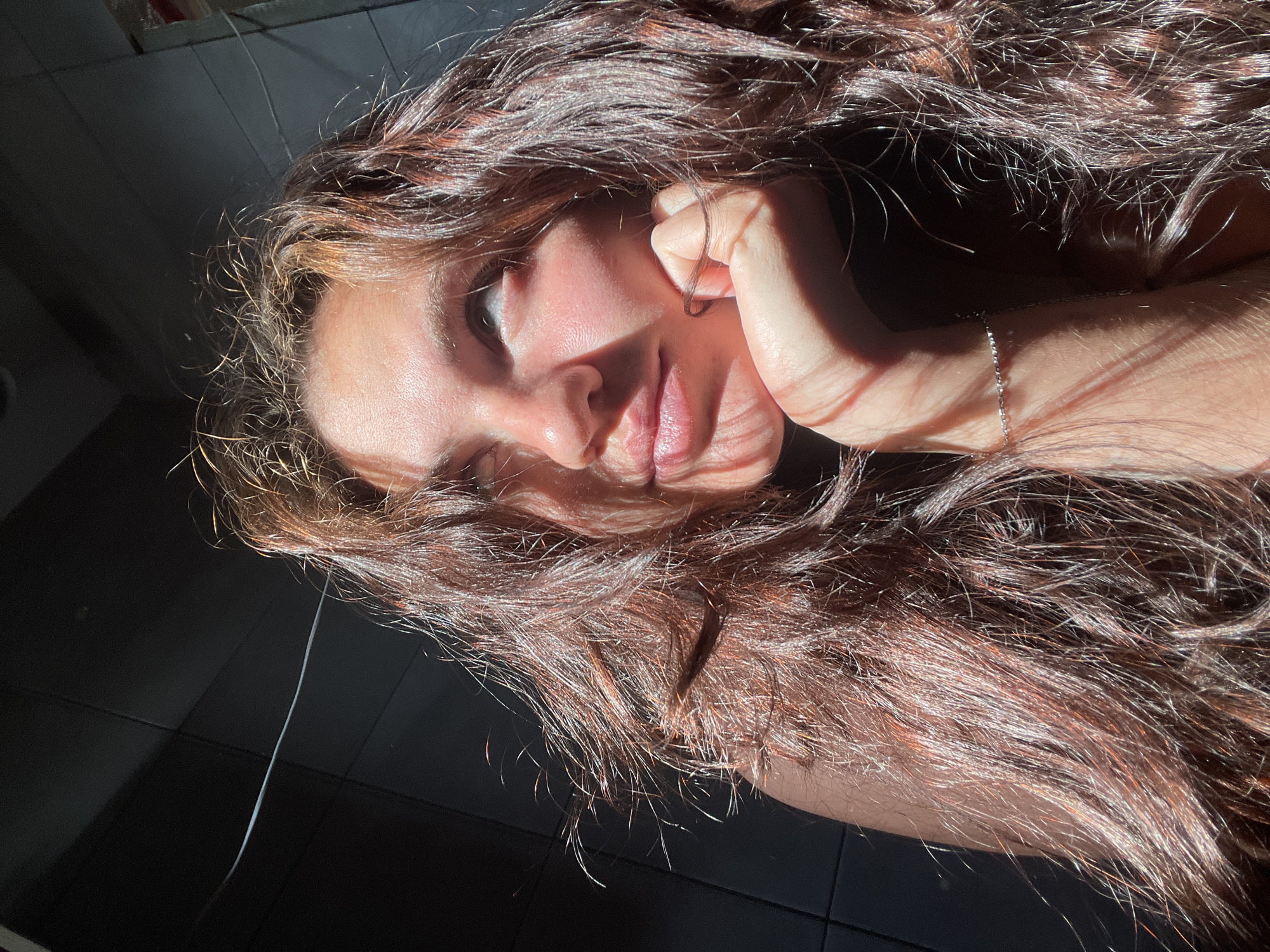 In the image, a young woman with long, wavy brown hair is captured in a close-up, looking upwards with a thoughtful expression. She rests her chin on her hand, and a delicate silver bracelet is visible on her wrist. Sunlight casts dramatic shadows across her face and hair, highlighting strands with a reddish hue. The background appears to be a dark, tiled wall. The scene is set in an indoor location, potentially a room or a studio, within the city of Mosmetalkonstruktsiya Ont, Russia.