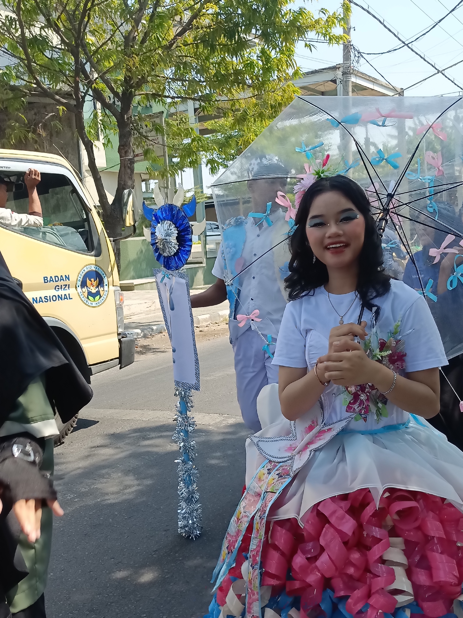 A young woman with dark hair, wearing blue eye shadow and a white t-shirt, smiles at the camera while holding a clear umbrella adorned with pink and blue ribbons. She is dressed in a white dress with a light blue sash and a skirt that features multiple layers of pink and blue ruffled fabric. She is also wearing a silver necklace and bracelets. In the background, a yellow van with "BADAN GIZI NASIONAL REPUBLIK INDONESIA" written on its side is visible. A person in a white uniform is standing next to the woman, holding a tall, decorated pole with blue and silver embellishments. Another person is partially visible in a black hijab on the left. The scene appears to be a parade or procession taking place on a paved street in Kediri City, Indonesia, with trees and buildings in the background. The sky is bright and sunny.