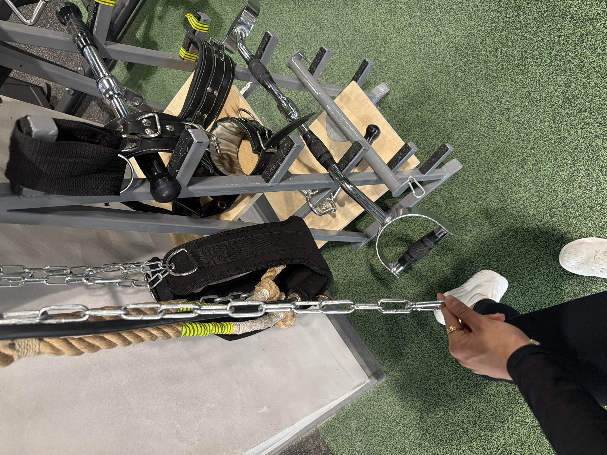 The image depicts a gym scene from an elevated perspective, focusing on gym equipment and a person interacting with a chain.
**People:**
* A person wearing black leggings, a long-sleeved black shirt, and white sneakers is visible from the mid-thigh down. The person's hand is holding onto a metal chain. A ring is visible on one of the fingers.
**Subjects/Objects:**
* Various gym accessories are present, including straps with buckles, T-bar handles, and a weightlifting belt with a chain attachment. A thick rope with yellow and green stripes is also attached to the chain.
* A rack or storage unit made of gray metal is present, holding some of the equipment.
**Environment:**
* The background is a green textured floor, indicative of a gym setting.
**Actions/Interactions:**
* The person is holding the metal chain, suggesting they may be setting up for or completing a workout.
* The equipment is organized on the rack and around the floor, showing active use within the gym space.
No information is available in the image to confirm if it is in Krasnoyarsk, Russia.