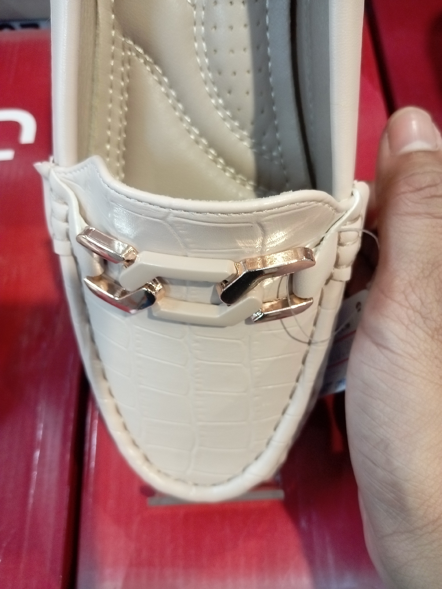 A close-up, low-angle, vertical shot shows the front of a cream-colored, alligator-embossed loafer. The shoe has a decorative metallic accent across the vamp, composed of two interlocking rectangular bars. A finger and thumb on the right side of the frame are gently holding the side of the shoe, with a white price tag dangling from a plastic string attached to the shoe. The shoe rests on a red surface, possibly a box or display. The scene is likely indoors, in a retail environment, such as a shoe store or department store in Batam, Indonesia.