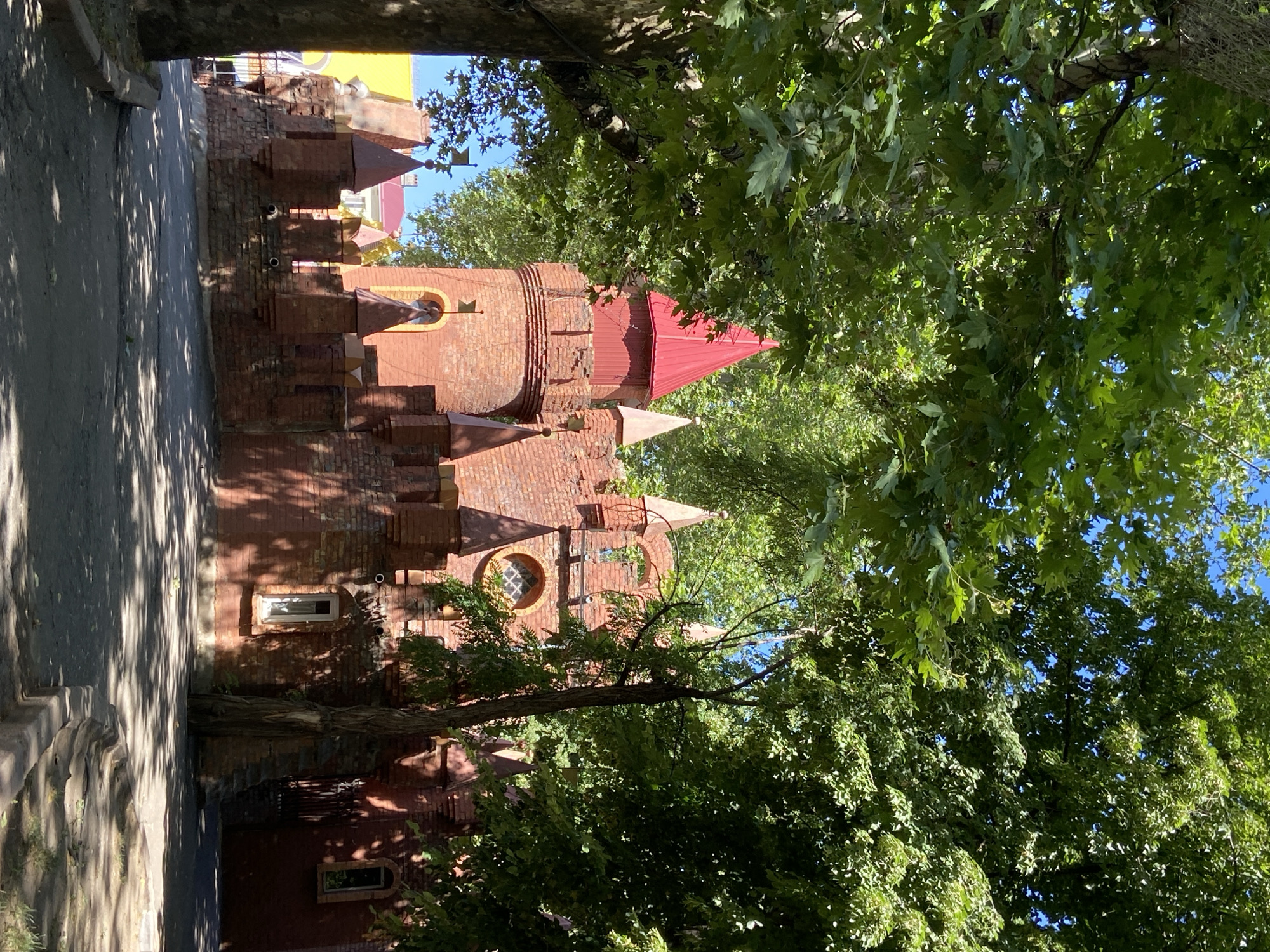 The image shows a red brick castle-like structure with several turrets and battlements, partially obscured by lush green trees. The sun casts shadows on the paved ground in front of the structure. The castle has a red conical roof on its tallest turret and smaller conical roofs on other turrets, some adorned with flags. There are arched windows and a circular window with intricate detailing. The environment appears to be outdoors, possibly in a park or amusement area, given the whimsical architecture. The sky is visible and appears clear blue. There are no people or animals visible in the scene.