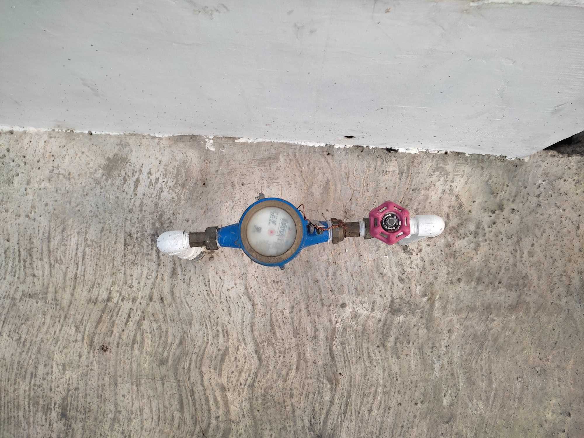 A blue and white water meter is horizontally mounted on pipes against a textured concrete wall. A pink valve is attached to the left side of the meter. The pipe is white on both ends and has brass fittings connecting it to the meter. The wall has a rough, uneven surface with visible lines and small dark specks. Below the wall is a lighter colored concrete surface. The image appears to be taken outdoors, possibly in a utility area.