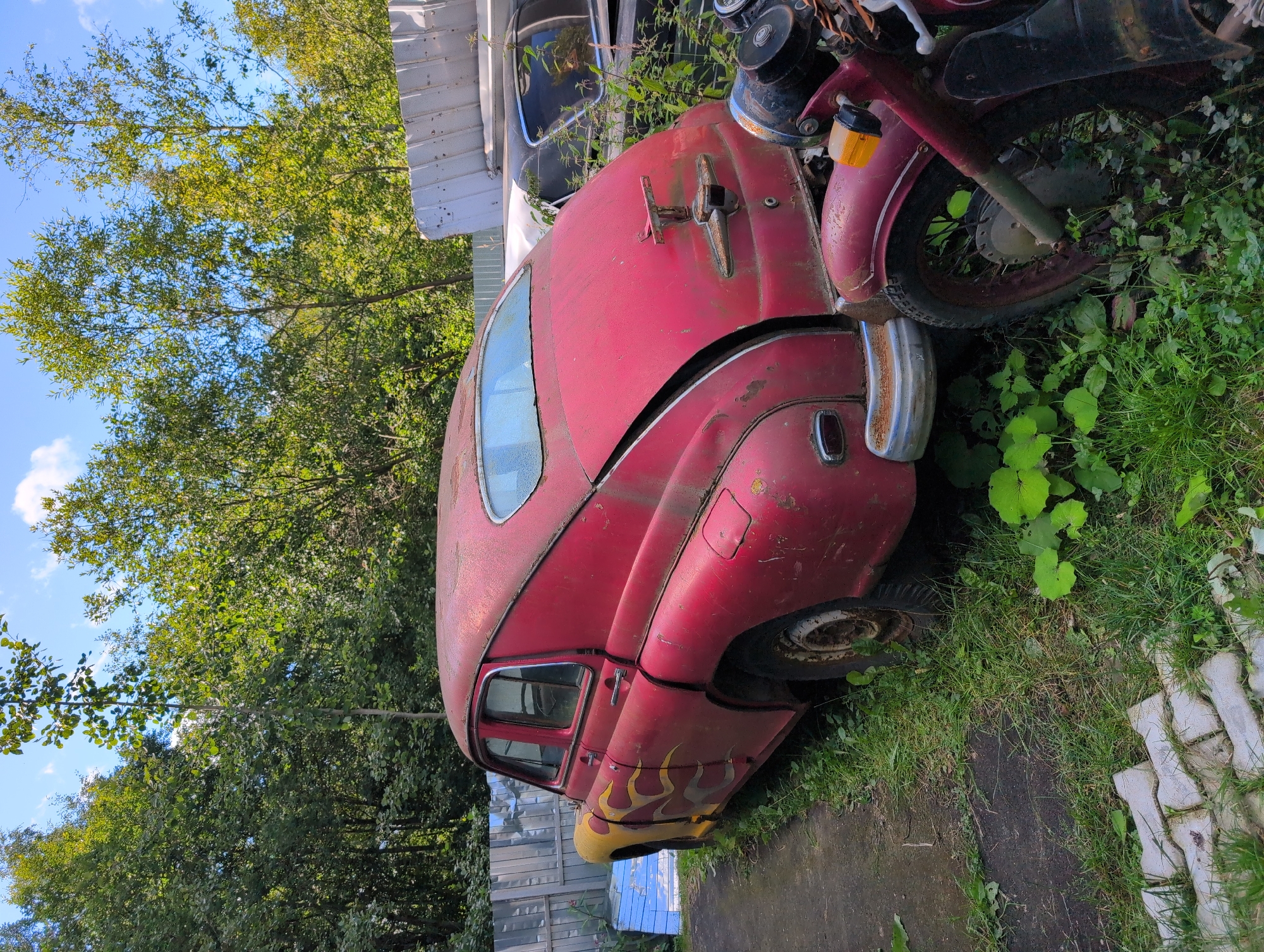 An aged, cherry-red four-door sedan with yellow flame decals on its side is parked on an unpaved surface adjacent to a chain-link fence. The car, appearing to be a GAZ-21 Volga, exhibits significant rust and wear, with a partially open trunk. In front of the sedan, partially obscuring its rear bumper, is a red motorcycle, also showing signs of age. The environment is overgrown with grass and various plants. Behind the fence, other vehicles and structures are visible, including what appears to be a white corrugated metal fence or building section and another car. The sky above is blue with scattered clouds. The location is identified as Novoe Devyatkino, Russia.