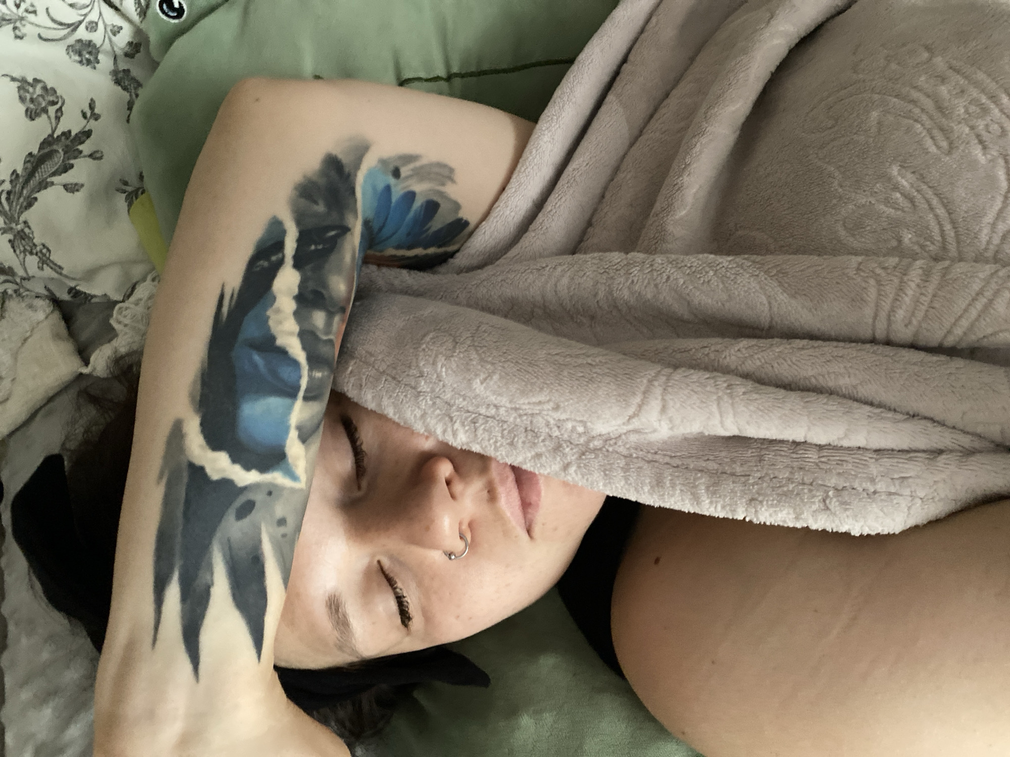 The image provides an overhead, close-up view of a person, likely a woman, sleeping or resting peacefully indoors. Her eyes are closed, and a soft, light gray blanket with an embossed floral pattern is pulled up, partially covering her face, revealing only her nose, mouth, and part of her cheek.
The person has visible freckles on her nose and cheeks, and a silver hoop nose ring adorns her left nostril. Long, dark eyelashes are prominent against her skin. Dark hair is visible around her head. A large, intricate tattoo covers her right arm. The tattoo design features shades of blue and black, depicting abstract elements that resemble a fragmented face or figure on the upper arm, along with sharp, dark, claw-like or feather-like shapes extending down the forearm. Lighter, torn-paper-like textures and softer blue, abstract shapes are also part of the design.
The person is lying on a light green pillow, with another white pillow featuring a dark floral pattern visible to the left. The lighting is soft and diffused, contributing to a tranquil and comfortable atmosphere. There is no visible text within the image.