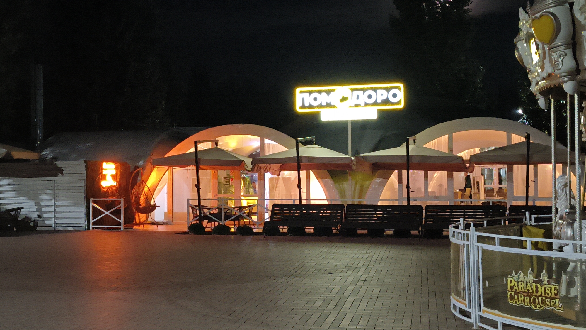 A nighttime outdoor scene in Mikhaylovka, Russia, features a cafe with illuminated arches and umbrellas, a neon sign that reads "ПОМДОРО" (Pomodoro) in Russian, and benches in front. To the right, a carousel with a sign reading "PARADISE CARROUSEL" is partially visible. The ground is paved with brick.