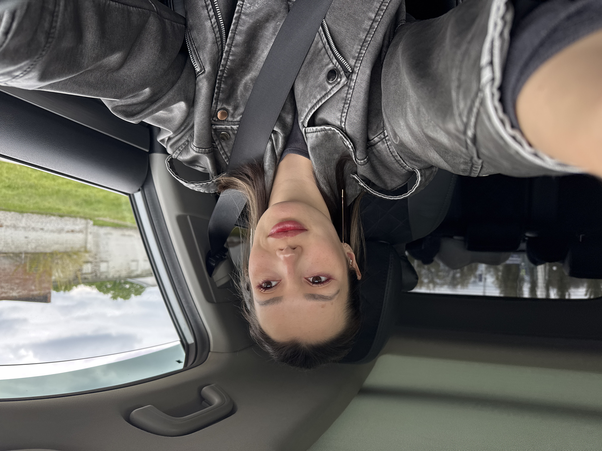 A woman is seated in a car, wearing a gray leather jacket and a seatbelt. The scene outside the car window shows a building and greenery under a cloudy sky. The image is oriented upside down.