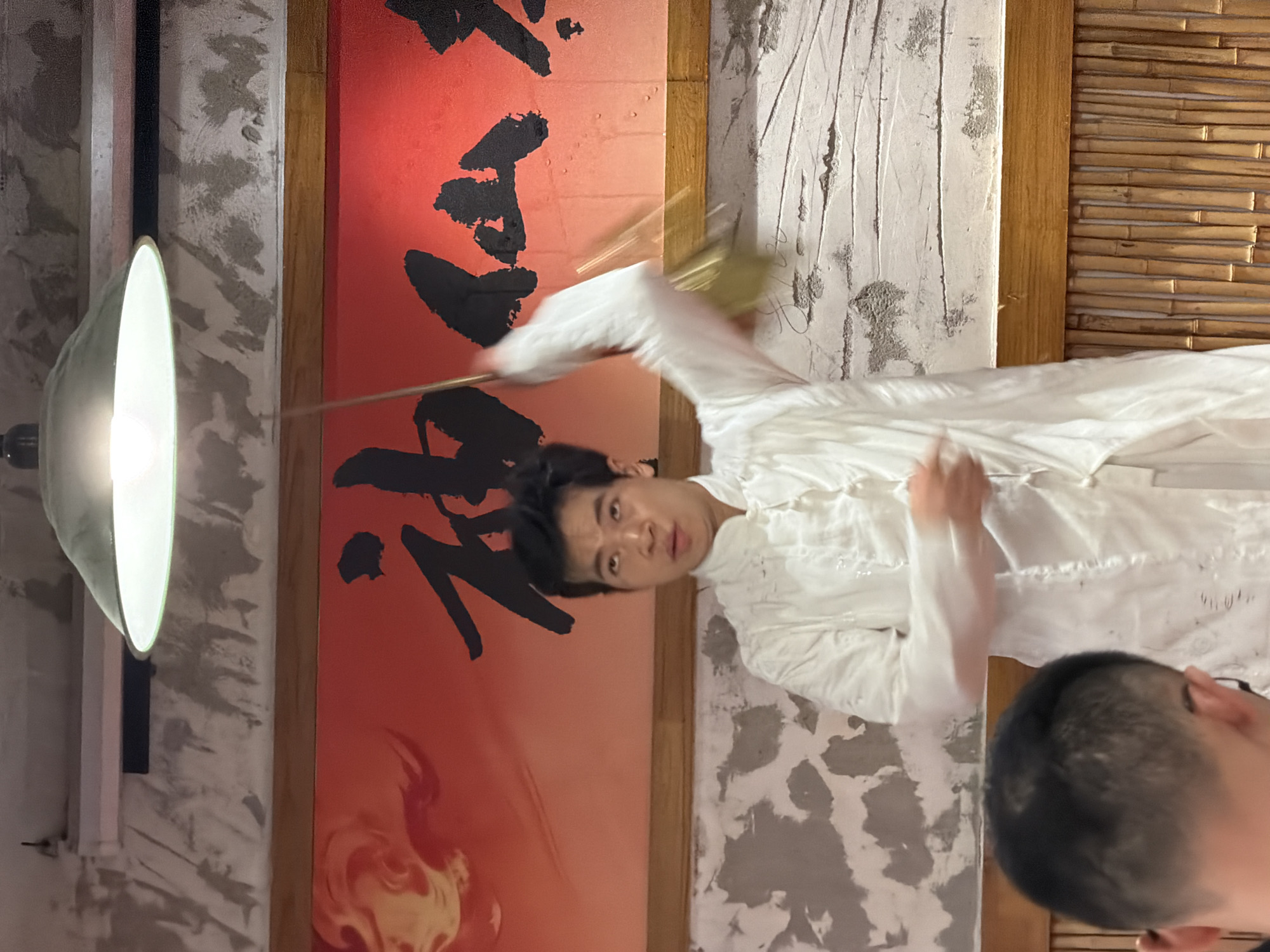 In a dimly lit room, a young man wearing a white traditional Chinese garment is performing a demonstration. He stands centrally, his right arm raised and blurred with motion as he wields a long, thin stick. A golden fan or similar object is attached to the end of the stick, also blurred by movement. His gaze is directed slightly upward and to his right, with his mouth open as if speaking or exhaling. Behind him, a large, vibrant red banner with black Chinese calligraphy and a swirling, fiery orange graphic is mounted on the wall. The wall above and below the banner has a textured, rough concrete finish, with horizontal wooden planks separating the different wall sections. A prominent metal lamp with a bright light fixture hangs from the ceiling, illuminating the scene. In the foreground, the back of another person's head is visible, with dark hair and a hint of glasses. The background features a decorative wall of vertical bamboo stalks. The image appears to be taken indoors, possibly in a cultural center or performance space in Yuzhong District, China.