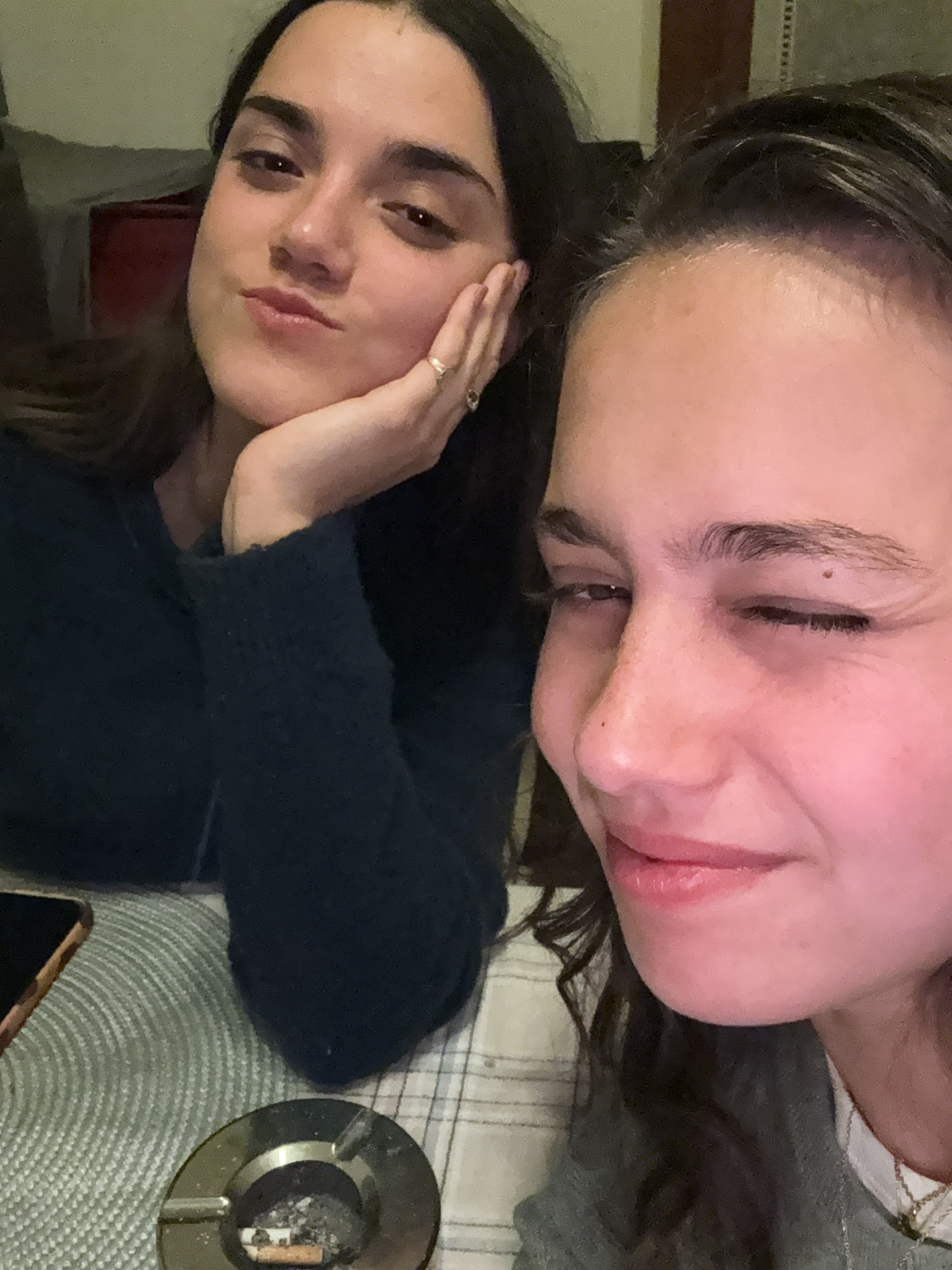 Two young women are posing indoors at a table. The woman on the left is resting her cheek on her hand and has dark hair, while the woman on the right is winking and has curly hair. An ashtray with cigarette butts and a phone are visible on the table, which is covered with a patterned tablecloth. The location cannot be identified as Milan, Italy without additional visual or contextual clues.