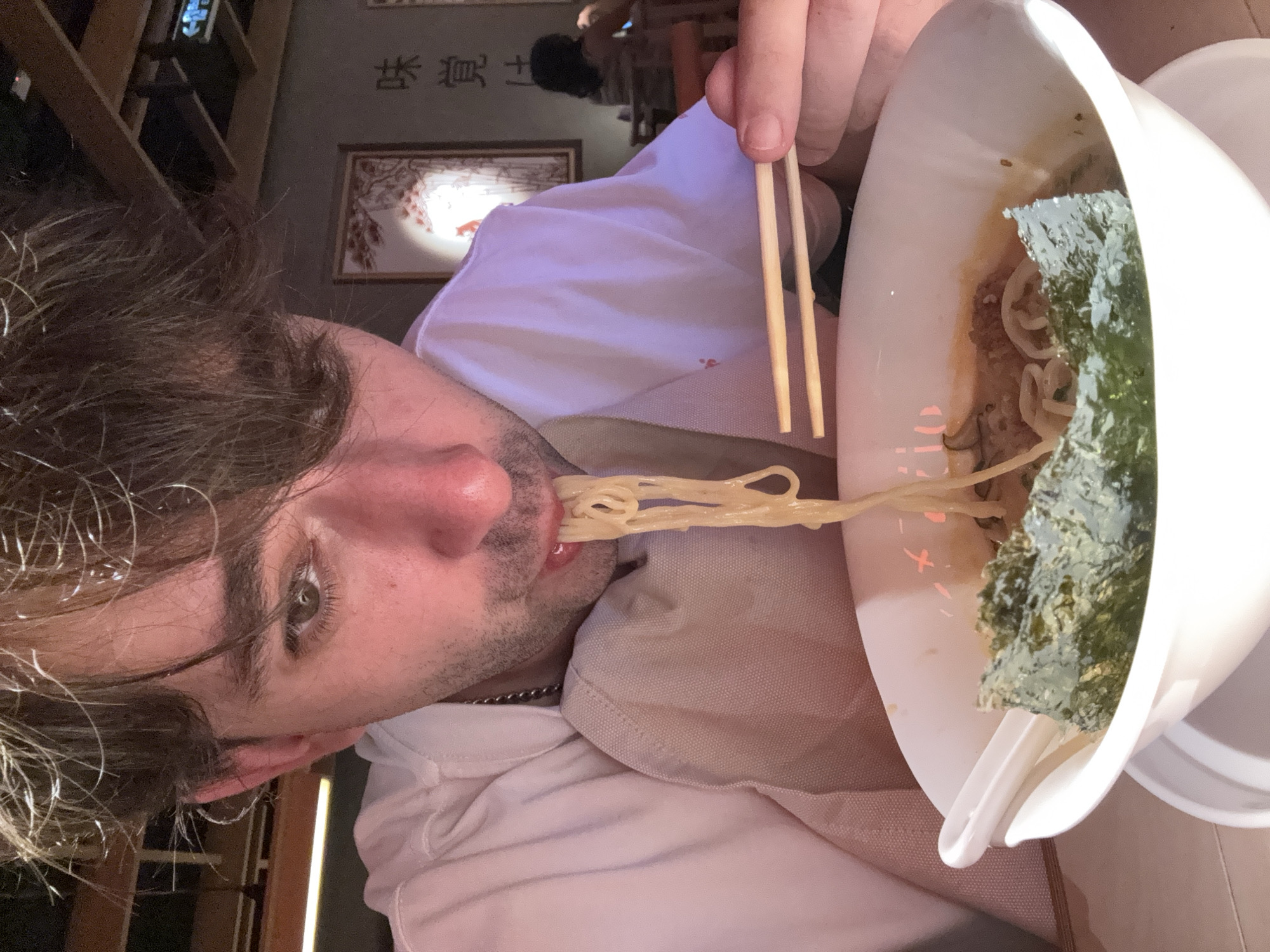 A medium close-up shot shows a person eating ramen at a table in what appears to be a restaurant. The person, with brown hair and light skin, is wearing a light-colored shirt and has a chain necklace. They are holding chopsticks to lift noodles from a white bowl, which also contains nori seaweed and other ingredients. The background shows a wall with Japanese-style artwork and other patrons seated at tables. The lighting is dim, suggesting an indoor evening setting. The scene does not provide any specific visual cues to identify the location as Moscow, Russia.
