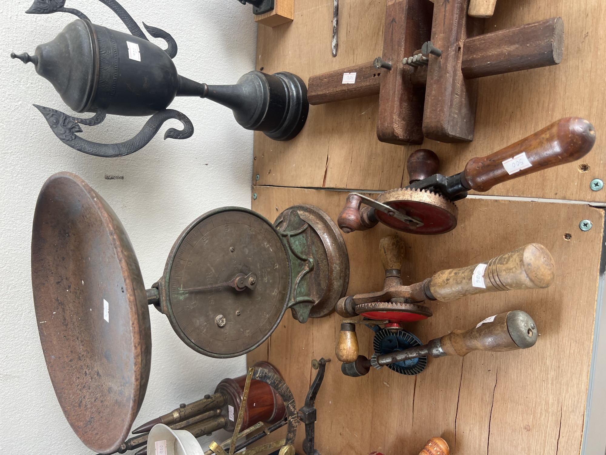 The image depicts a collection of antique tools and decorative items displayed on a wooden surface, likely in a shop or market stall in Tambon Phraek Ha, Thailand. In the foreground, two hand drills with wooden handles and metal components are positioned horizontally. To the right of the drills are several rectangular wooden blocks, some with metal screws and a drill bit. Behind the drills, a vintage scale with a large, rusty metal pan and a circular gauge is prominently displayed. To the right of the scale, a dark, ornate trophy with decorative handles and a conical lid stands on a black base. In the background, a white wall is visible, and further to the left, partially obscured items including a white bowl and brass-colored equipment are present. The lighting suggests an indoor setting, possibly with natural light from a window or artificial light sources. The overall impression is one of an organized display of old, functional, and decorative objects.