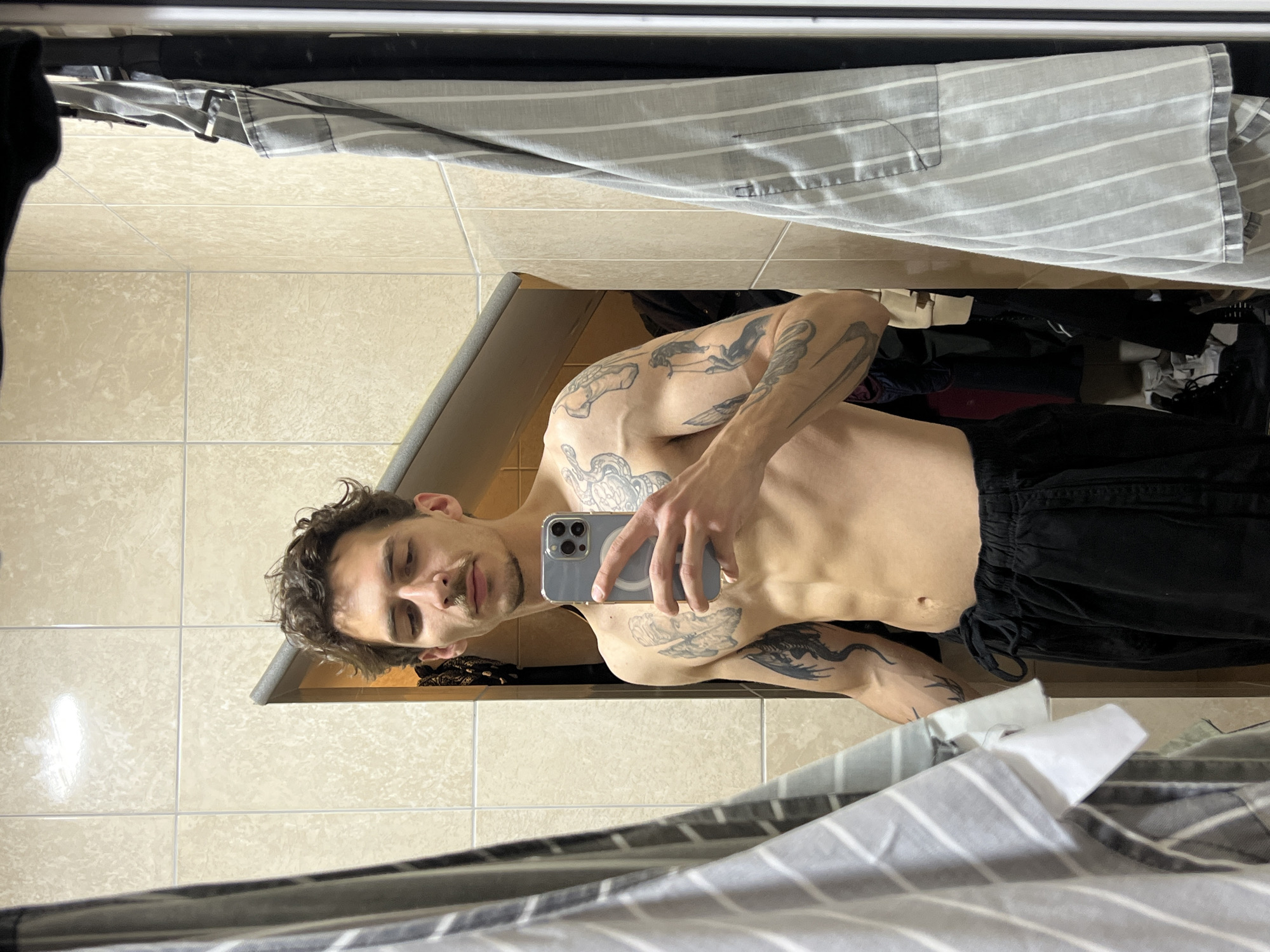 A shirtless man with tattoos is taking a selfie in a tiled room with clothes hanging in the background. The location is not discernible as Budva, Montenegro from the provided visual information.
