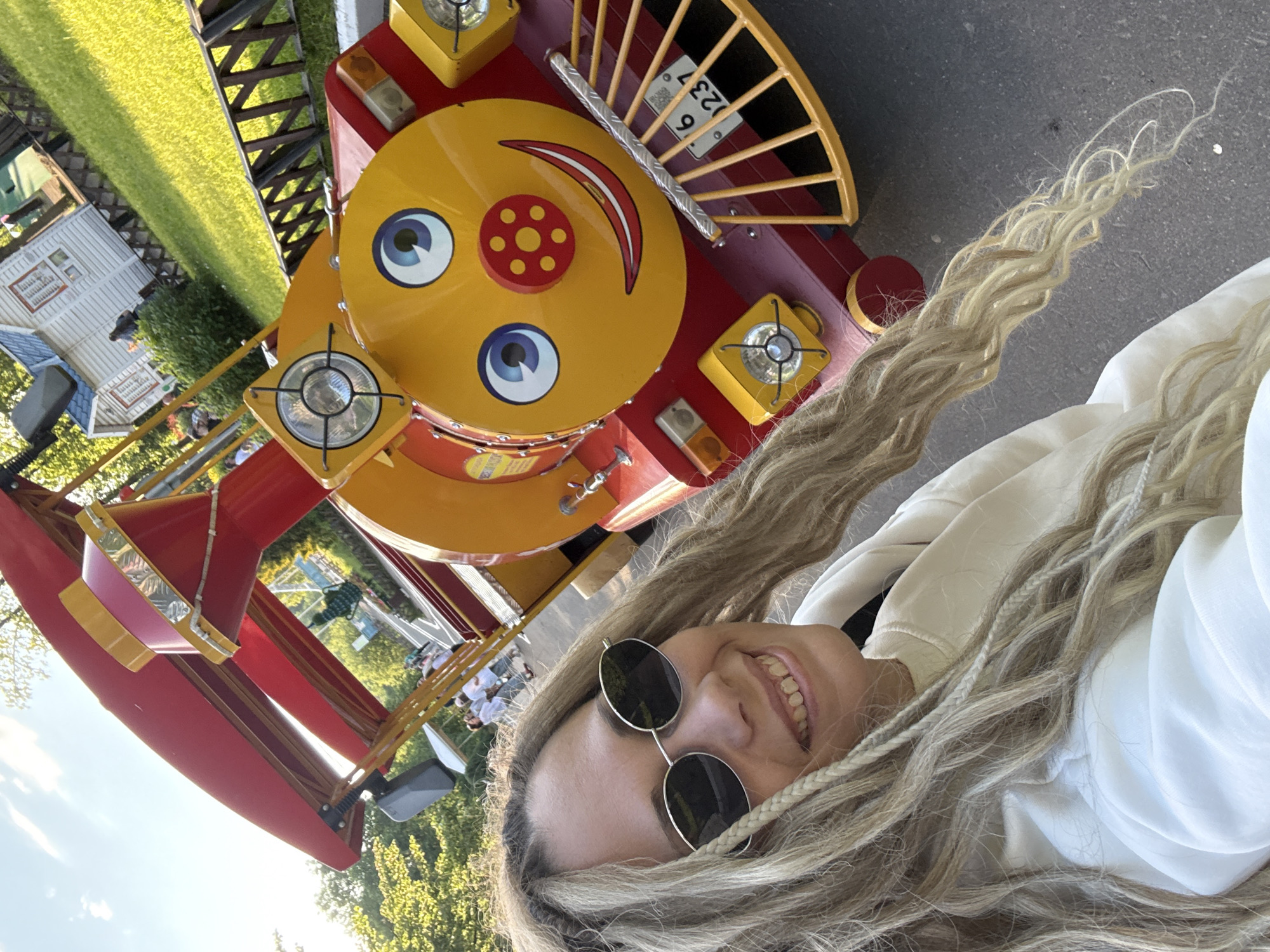 The image captures a cheerful outdoor scene, likely at an amusement or children's park in Заволжье, Russia. The primary subject is a young person taking a selfie, positioned in the foreground and smiling broadly towards the camera. They have long, light blonde hair styled in dreadlocks or braids, wear round dark sunglasses, and are dressed in a white hoodie.
Behind the person is a vibrant, miniature train engine, which appears to be part of a ride. The train is primarily red and yellow, featuring a large, cartoonish smiling face on its front, complete with wide blue eyes, a red nose, and a curved red smile. It has square headlights on either side of its face. A small sign on the train displays the number "6237".
The background reveals a sunny day with clear skies and some clouds. There is lush green grass and trees, along with several small, white, dollhouse-like buildings that suggest a miniature village or an elaborate play area within the park. The overall atmosphere is joyful and playful. The image itself is rotated 90 degrees counter-clockwise.
