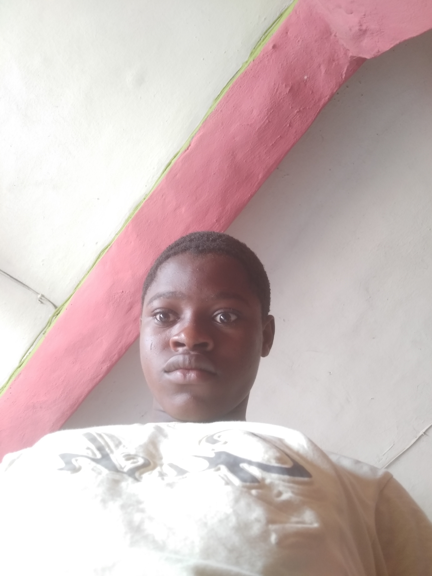 A person is shown in a close-up, low-angle shot. The person is facing forward with a neutral expression. They are wearing a light-colored shirt with a design on it.
The background consists of a white wall with a pink-painted architectural feature, possibly a beam or a wall edge, running diagonally across the upper part of the frame. The location is Shangisha, Nigeria.