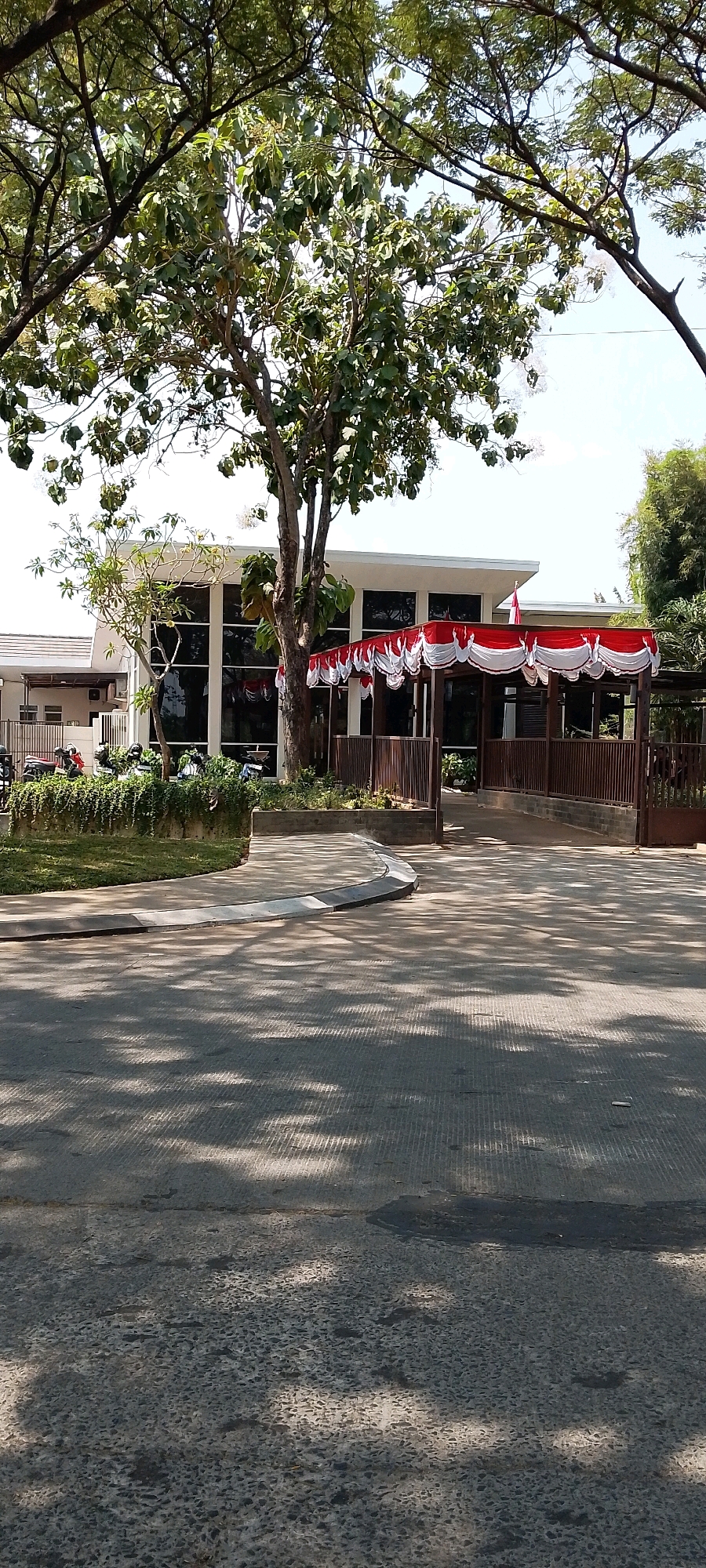 The image depicts the exterior of a building with a large, modern facade featuring prominent glass windows. In front of the building, a covered walkway is adorned with red and white fabric decorations, resembling banners. A large tree with lush green foliage stands to the left of the walkway, casting shadows on the concrete pathway in the foreground. Several motorcycles are parked to the left of the building, behind a low fence and an area of greenery. The overall scene suggests an outdoor setting, possibly a public building or institution, in Satriamekar, Indonesia, as indicated by the red and white decorations which are characteristic of Indonesian national colors, often displayed during national holidays or significant events. The environment appears to be sunny, with the shadows suggesting it is midday. There are no people clearly visible or engaged in any discernible actions.