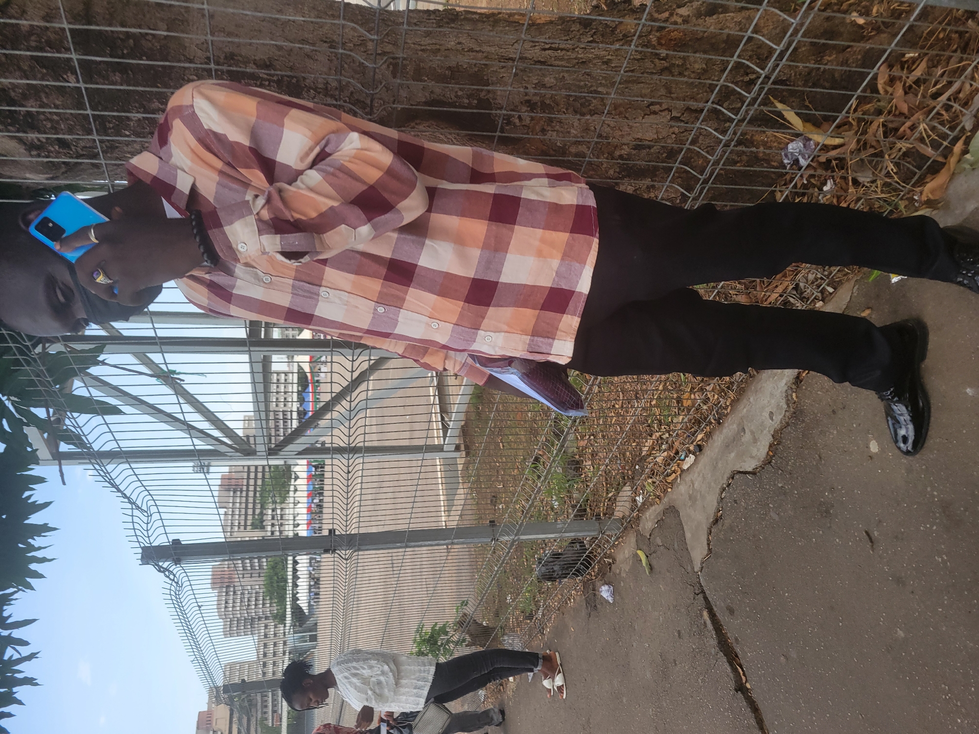 The image shows a man in a patterned shirt, dark pants, and black shoes talking on a blue phone, standing near a metal fence. There is a tree and some foliage behind the fence, and a sidewalk with cracked pavement in the foreground. In the background, buildings are visible in the city. The general location is likely Abuja, Nigeria. Another person is visible further in the background, standing near the fence as well.