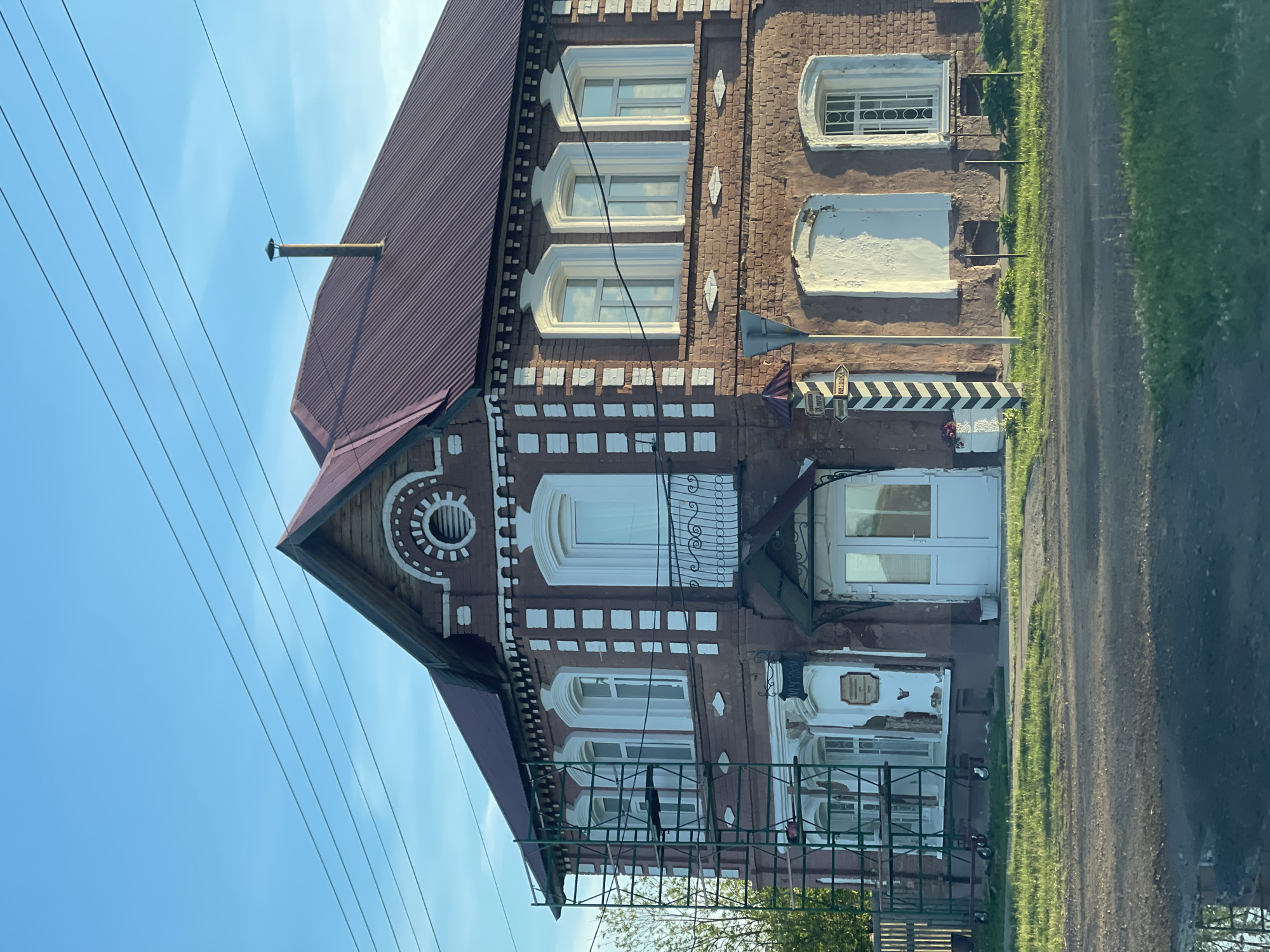 The image shows a two-story brick building with a dark red, corrugated metal roof. The building has several windows on both floors, many with white trim. Some of the windows are arched. The facade of the building features white decorative elements around the windows and along the roofline. Scaffolding is visible on the left side of the building. There is a balcony above the entrance, which has white double doors. A chimney protrudes from the roof. A signpost with black and white stripes stands in front of the building. The building is situated next to a grassy area and a road, with power lines stretching across the sky. The sky is clear and blue.
