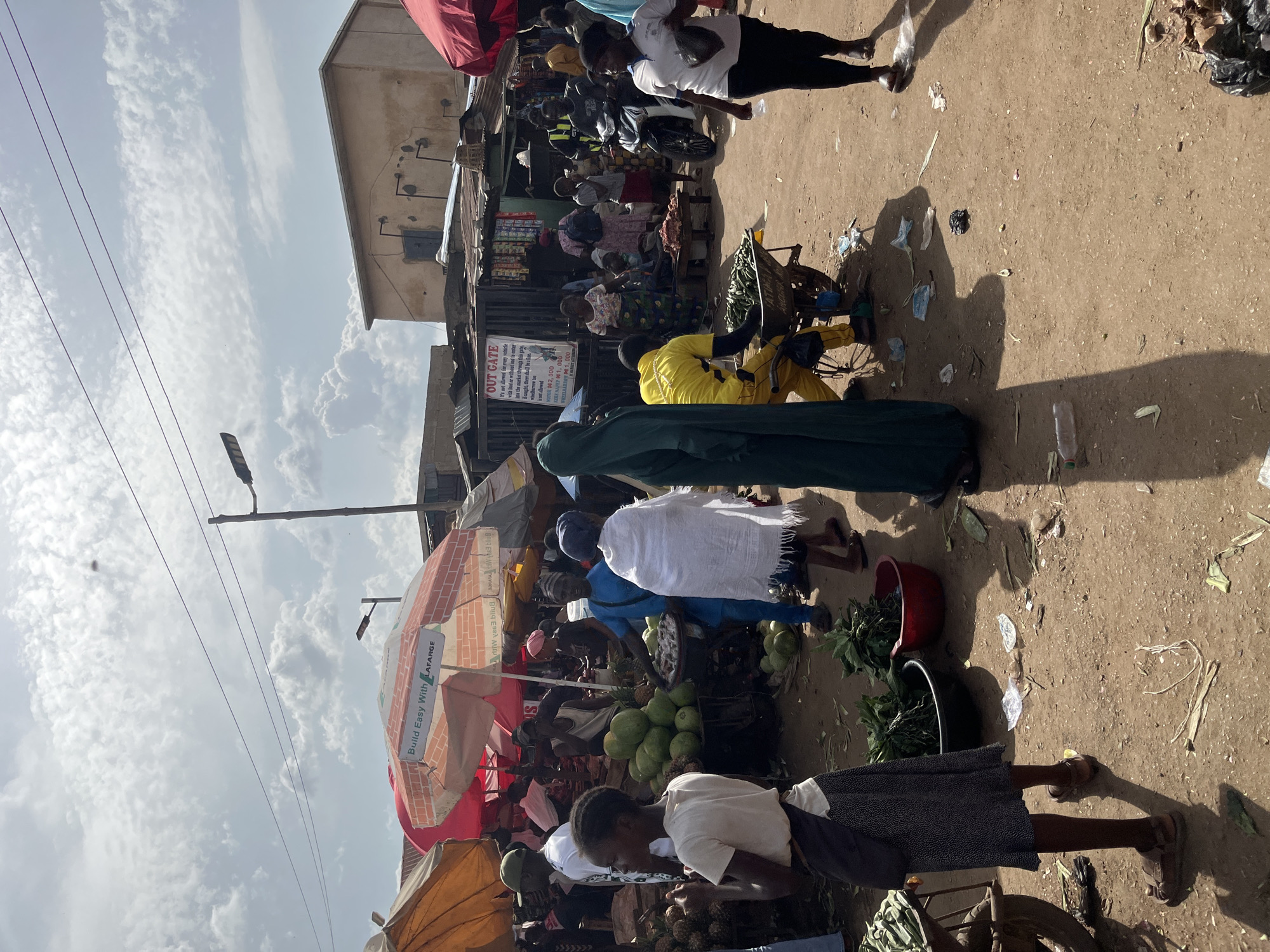 This is a vertical, eye-level shot of a bustling outdoor market in Rafin Par, Nigeria. The sun is high in the sky, indicated by the strong shadows and the bright, cloudy weather. The scene is filled with people, likely vendors and shoppers, engaged in commerce. Several market stalls are visible, covered by colorful awnings, including a red and white umbrella with the Lafarge logo, and an orange one.
The ground is a dusty, reddish-brown dirt, littered with some debris. In the foreground, a young woman in a white shirt and dark patterned skirt is looking down at produce, possibly buying or selling. Behind her, a person in a blue shirt carries a basket of green vegetables on their head. Further back, a person in a bright yellow shirt is on a bicycle, and another individual in a dark green robe walks past. Various fruits and vegetables, including what appear to be pineapples and other round produce, are displayed for sale.
In the mid-ground, several stalls are lined up with goods. There's a sign that reads "OUT GATE" with rules about vehicles and fines, suggesting this is an entrance or exit to a market area. A few motorcycles are parked nearby. The overall impression is one of a lively, everyday market scene.