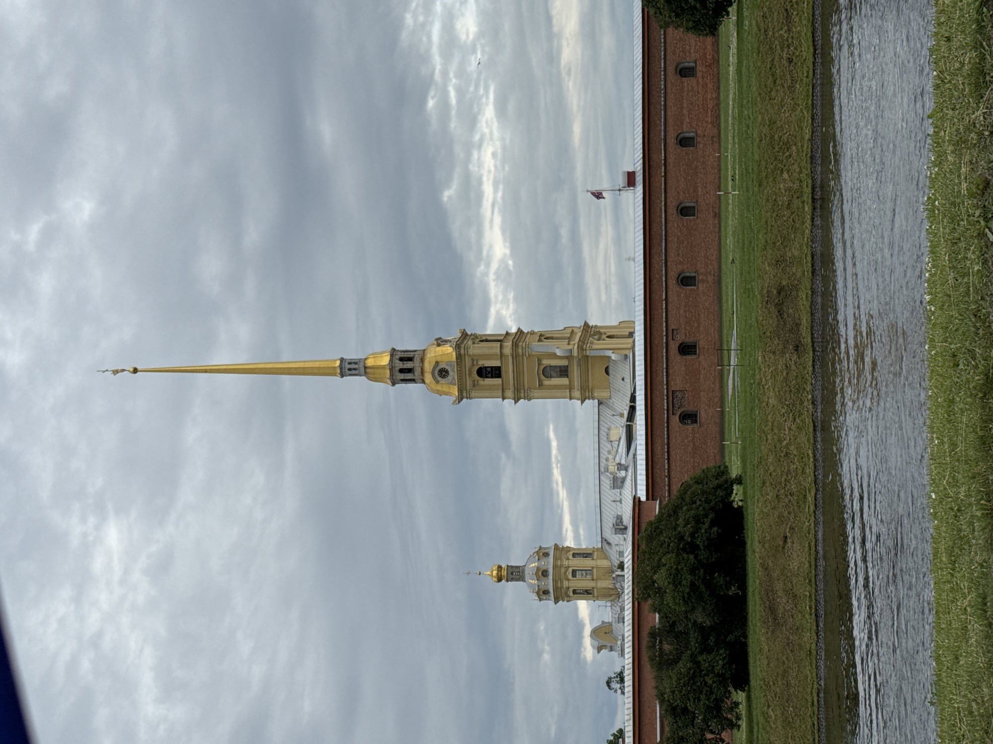This image depicts the Peter and Paul Fortress in Saint Petersburg, Russia. The fortress, characterized by its red brick walls and numerous arched windows, stretches across the midground. Dominating the skyline are two prominent church spires, both capped with golden domes. The tallest and most central spire is elongated and tapers to a sharp point, topped with a gilded angel. To its left, a shorter, broader spire with a similar golden dome is visible. The sky is overcast with heavy, gray clouds, casting a muted light over the scene. In the foreground, a body of water, likely a moat or canal, reflects the cloudy sky. Lush green grass covers the banks of the water and extends up to the fortress walls. A dense, dark green tree is situated to the left of the fortress walls. A single flagpole with a red and white flag is visible on top of the fortress wall, to the right of the main spire. The overall atmosphere is somber and somewhat dramatic due to the cloudy weather.