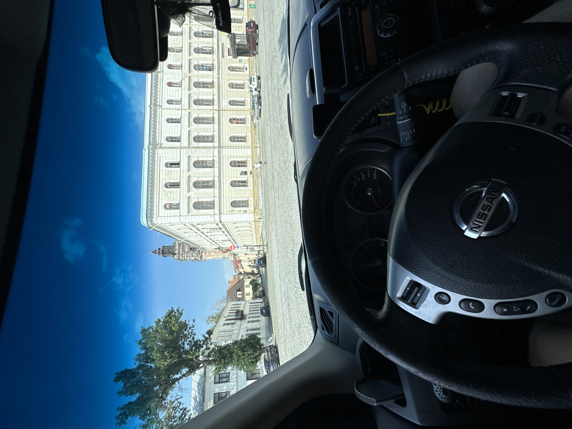 The image is taken from inside a Nissan car, looking out through the windshield. The car is parked on a cobblestone street in front of a large, ornate building with a light-colored facade and arched windows. To the left of the building, there is a tall, narrow tower with a dark spire. The sky is blue with some white clouds. The foreground shows the car's steering wheel, dashboard, and part of the interior. A few cars are parked on the street, and one person is walking in the distance. The scene appears to be a historical city center. Based on the architecture and the presence of the tower, the location is likely the main square in Kamianets-Podilskyi, Ukraine, with the building being the Town Hall or a similar historical structure.