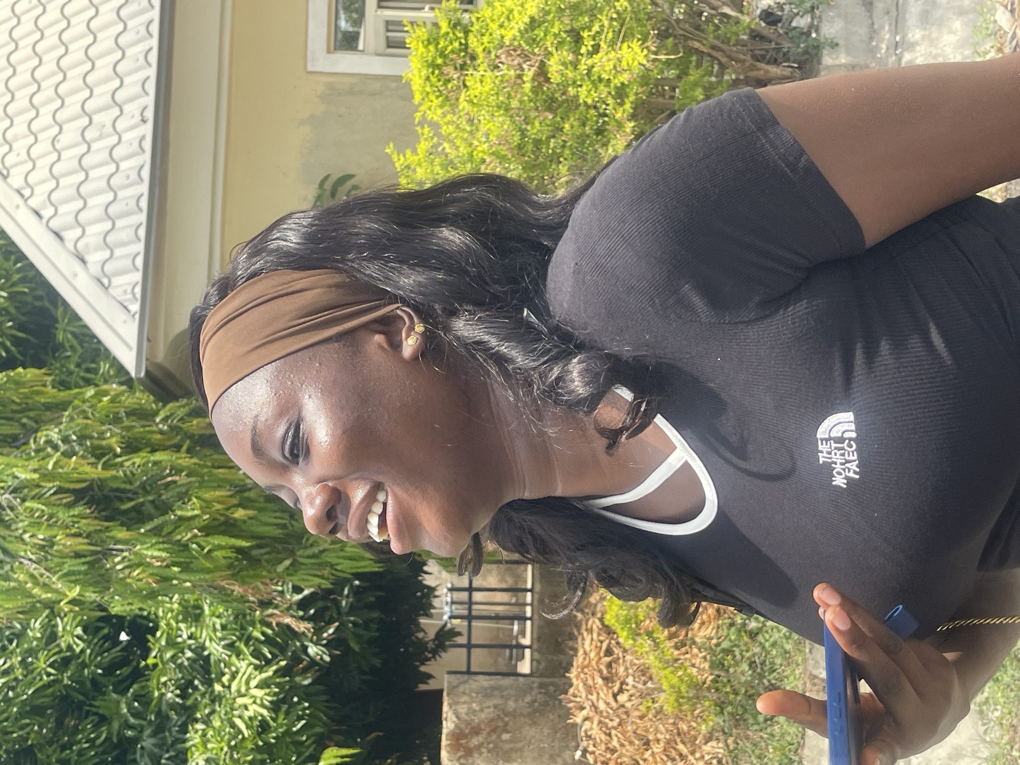 A vibrant, mid-shot outdoor portrait captures a young woman in bright daylight, rotated 90 degrees counter-clockwise from a typical upright orientation. The subject, a woman with dark skin, long, wavy dark hair, and a cheerful expression, is smiling broadly, revealing her teeth. She wears a brown headband that pushes her hair back from her face, and small gold stud earrings are visible.
She is dressed in a black, ribbed short-sleeved t-shirt with a white trim around the neck and shoulder area. On the left side of her chest, a white logo is visible, which appears to be a stylized or mirrored version of "THE NORTH FACE," reading "THE NORHT FAEC." She is holding a blue smartphone in her left hand, and a gold chain bracelet is partially visible on her wrist.
The background suggests an outdoor setting in Abuja, Nigeria, under a bright, sunny sky, evidenced by strong highlights on her skin, which shows a slight sheen consistent with warmth. Behind her, a light yellow building with a white-framed window and a distinctive white roof with wavy tiles can be seen. The scene is also rich with green foliage, including bushes and trees, along with areas of dried leaves and visible ground or concrete. The overall impression is one of happiness and casual outdoor activity.