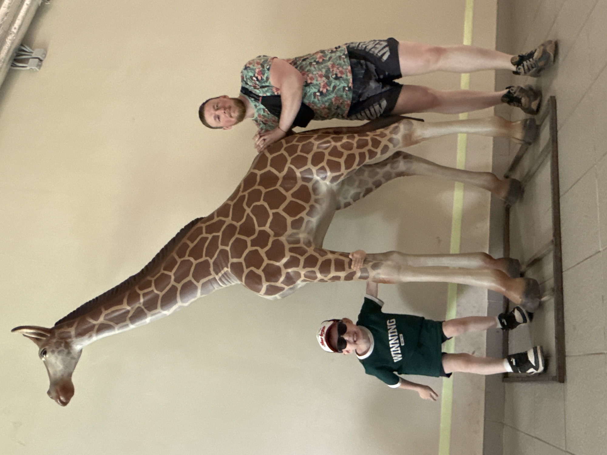 A young boy wearing a green "WINNING" t-shirt, a red and white baseball cap, and sunglasses stands with his left hand on the leg of a life-size giraffe statue. Next to him, a man with a beard wearing a floral print t-shirt, black shorts with grey accents, and a black fanny pack, leans against the giraffe statue with his hands resting on it. The two subjects are positioned in front of a light-colored wall, with the floor appearing to be tiled. The giraffe statue is standing on a metal base.