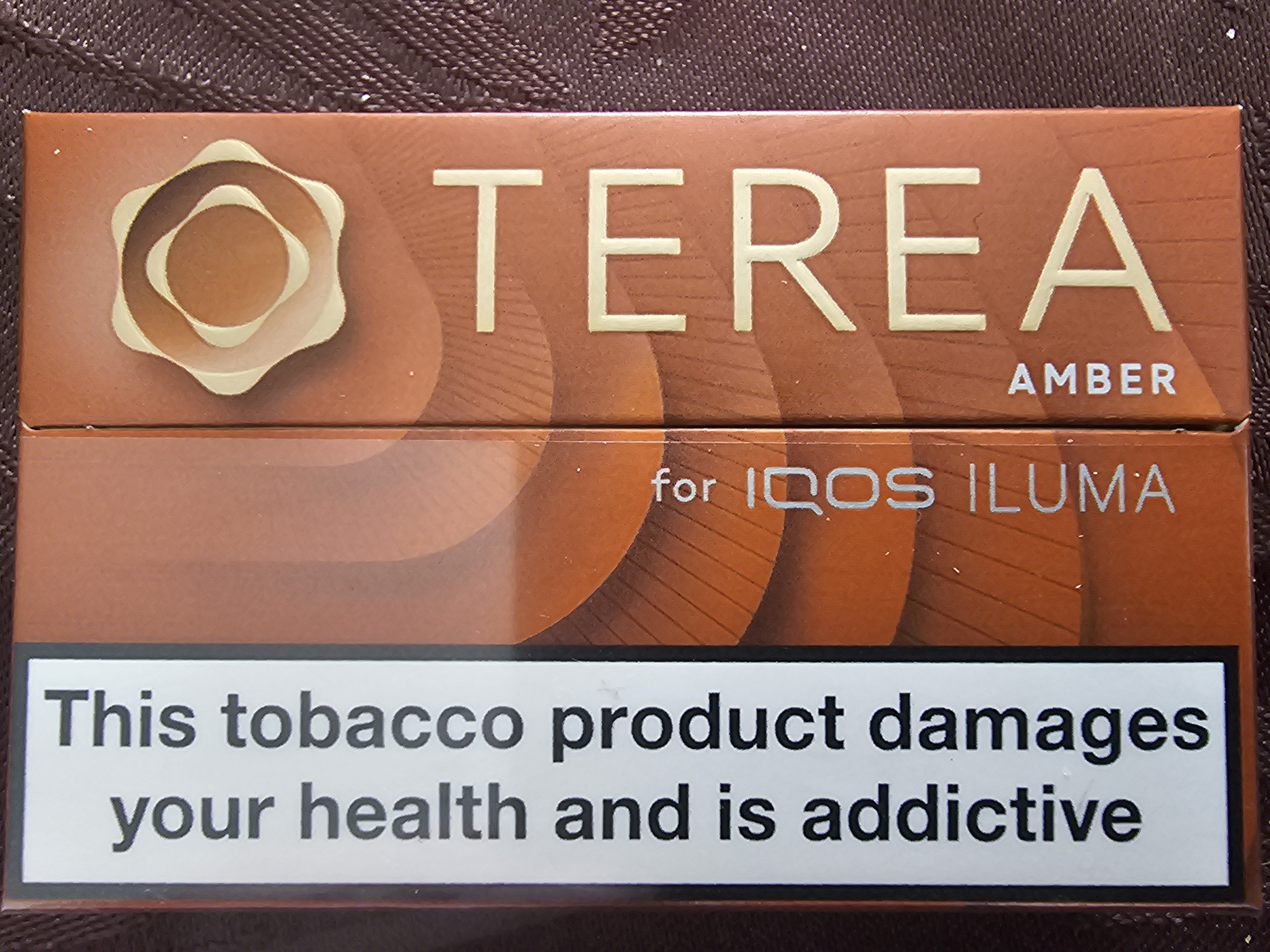 The image presents a close-up, eye-level view of a TEREA tobacco product box, designed for use with an IQOS ILUMA device, resting on a textured, dark brown surface. The box itself is a matte amber-brown color.
The top portion of the box features the brand name "TEREA" in large, light beige, embossed capital letters. To the left of "TEREA" is a distinct logo, composed of concentric, wavy, and irregular oval shapes in shades of light beige and amber. Below the main brand name, the word "AMBER" is visible in smaller capital letters, indicating the flavor or variant.
A horizontal line separates the upper and lower sections of the box. Centered below this line are the words "for IQOS ILUMA" in a light beige font, specifying the compatible device.
The most prominent feature of the bottom half of the box is a rectangular white warning label with a black border. This label contains bold black text stating: "This tobacco product damages your health and is addictive."
The background is a blurred, dark brown, textured surface, possibly fabric, emphasizing the product itself. The lighting is even, suggesting an indoor setting. There are no people or specific activities depicted. The clear English text on the packaging and warning label is consistent with the provided Holywell, United Kingdom location context.