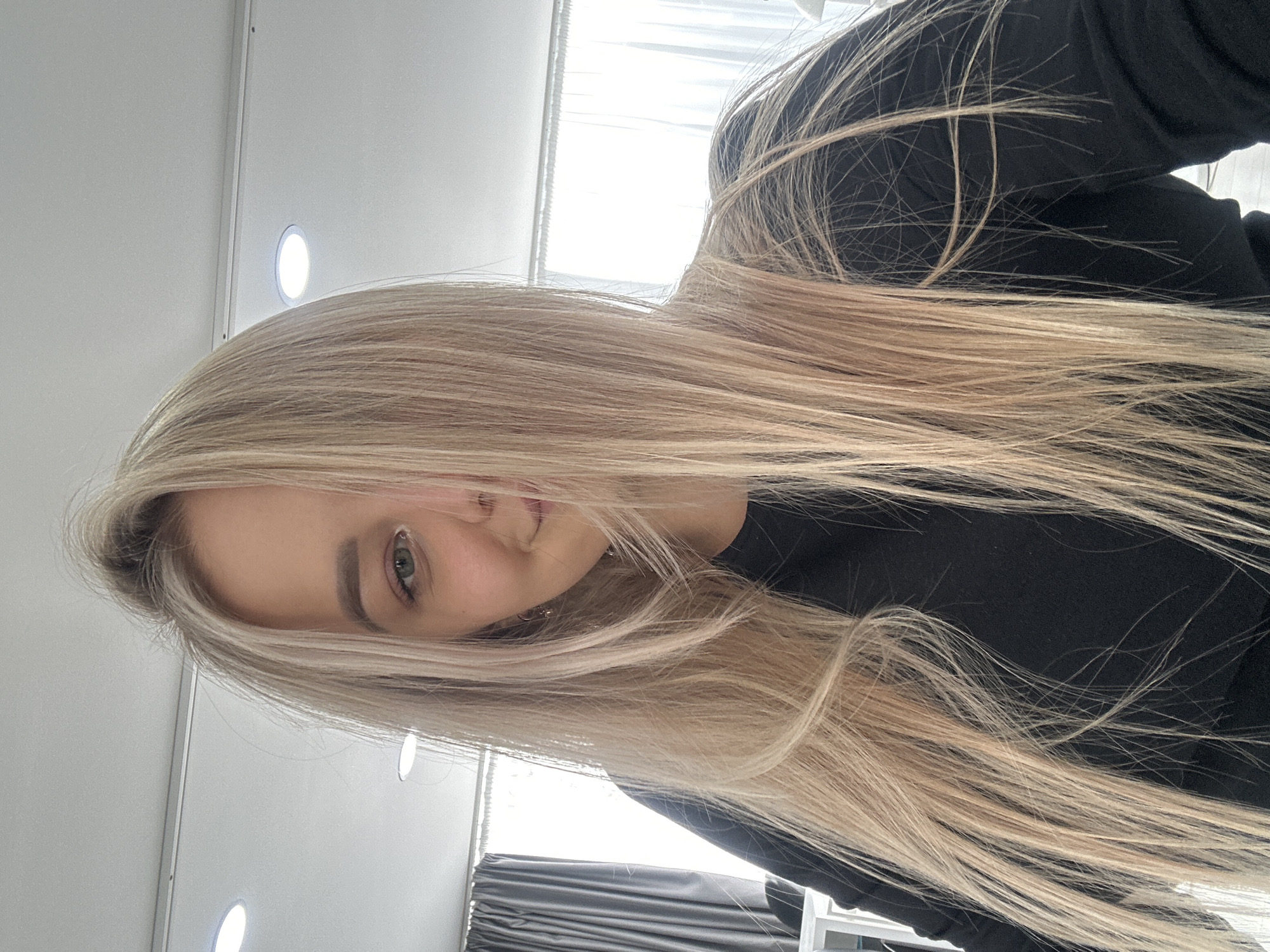 A blonde woman with fair skin and green eyes is angled towards the camera. She is wearing a black top. Light reflects on her face from a source above. The background shows a wall with built-in lights, a window with blinds, and a glimpse of what appears to be furniture or equipment. Based on the image's origin, the location is likely in Budva, Montenegro.