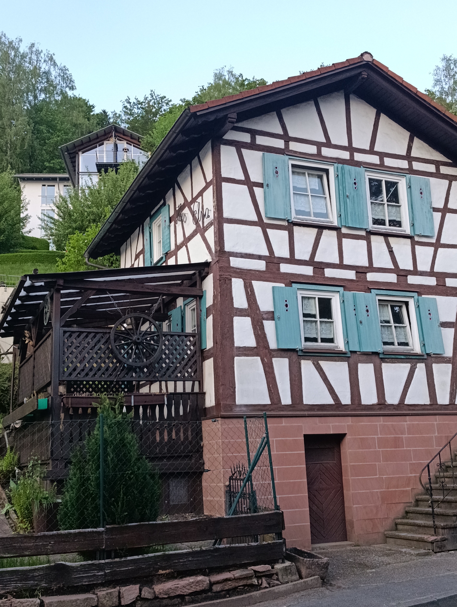 The image shows a half-timbered house with white walls and brown wooden beams, located in an unidentified city. The house has two stories and a reddish-brown brick base with a dark wooden door. Windows on both floors have blue shutters. A covered porch with a decorative wooden wheel hanging from the roof is attached to the side of the house. A chain-link fence runs along the front of the property, and wooden stairs lead up to the side of the house. In the background, there is a modern-style house with glass walls, partially obscured by trees. The sky is pale and overcast.