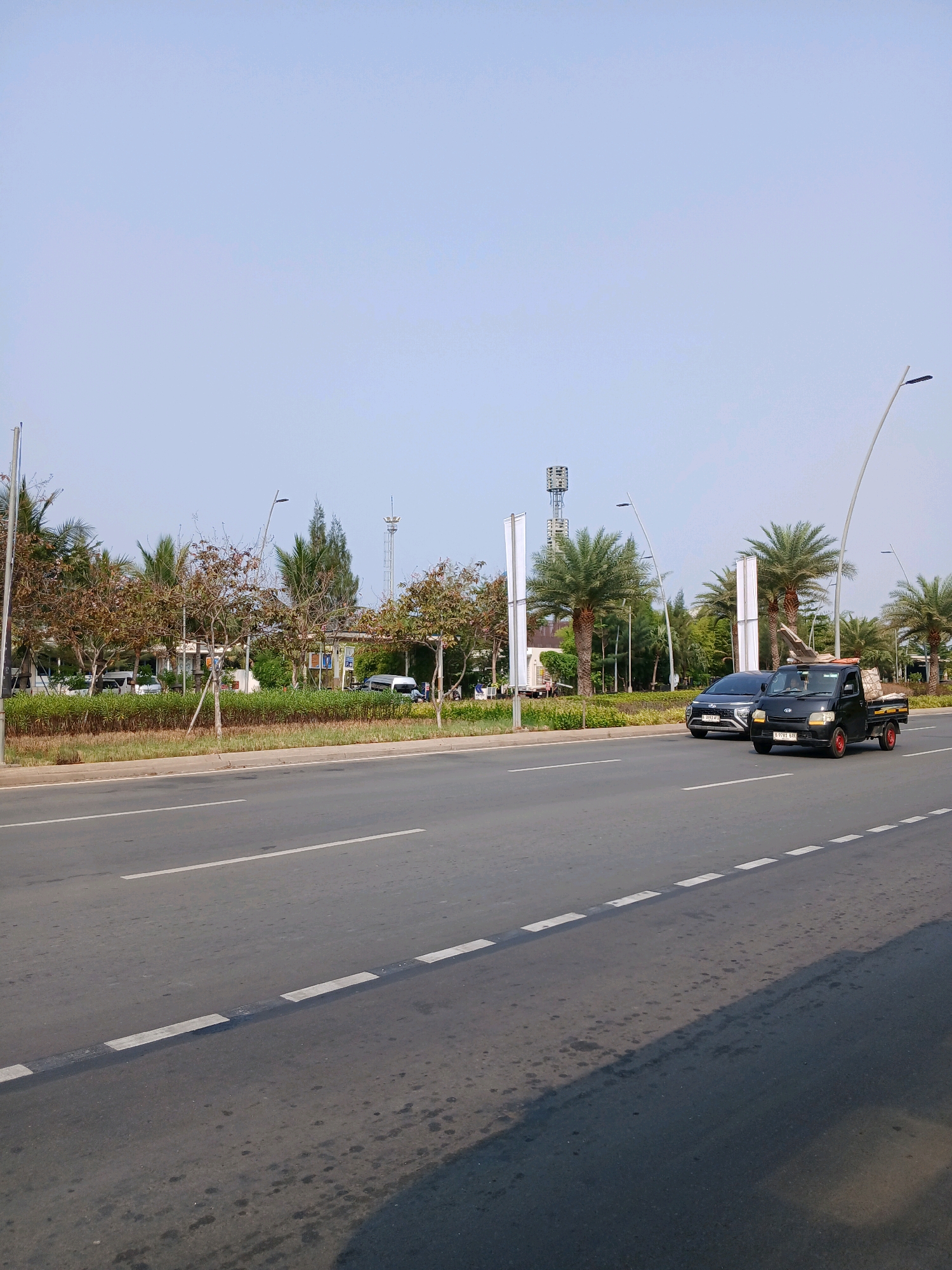 The image depicts a street scene in Kosambi Barat, Indonesia. A black pickup truck, carrying what appears to be cargo, drives in the right lane of a paved road. Next to it, a silver car travels in the left lane. The road is marked with white dashed lines. The background features a median strip with low green bushes and several trees, including palm trees. In the distance, there are some buildings and a tall telecommunications tower. The sky is clear and blue.