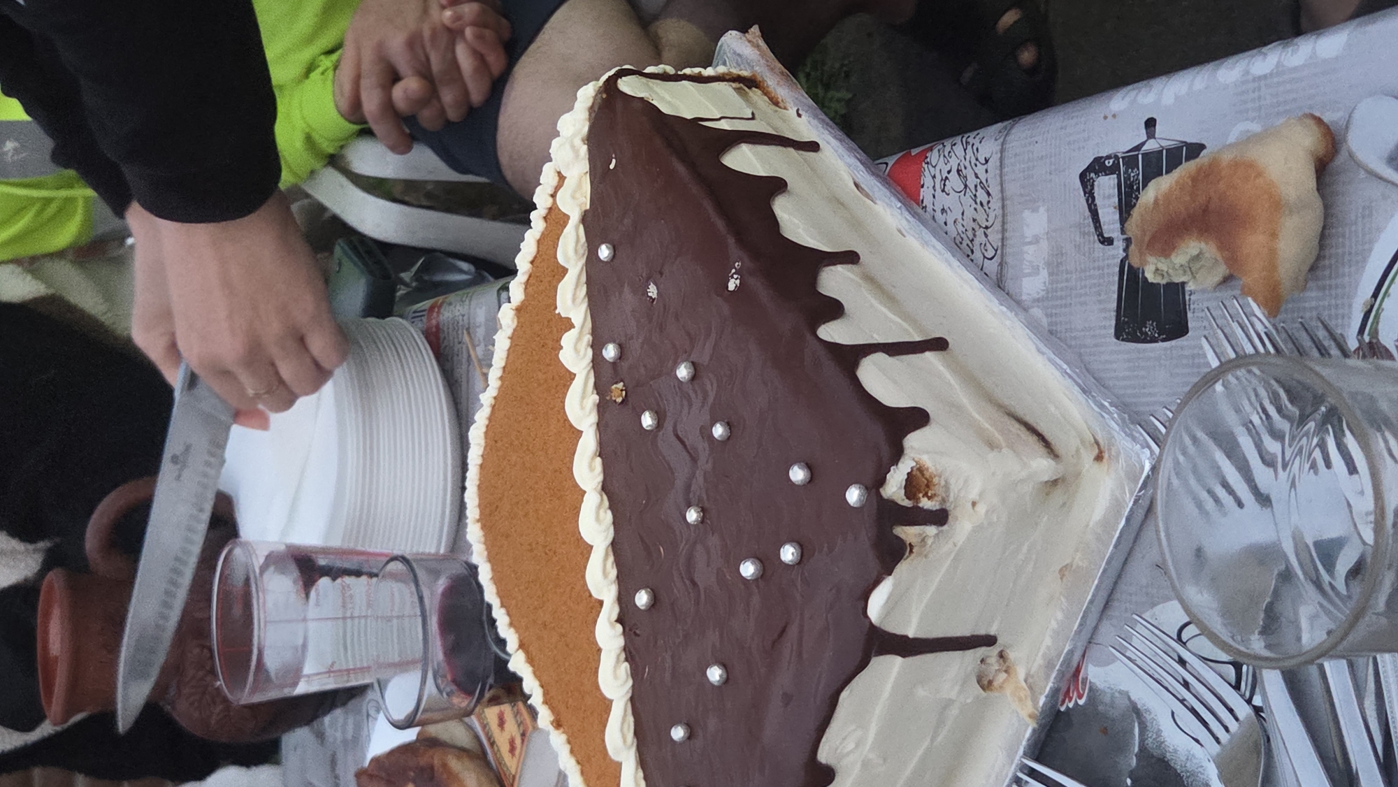 An overhead, medium shot captures a table setting for a dessert event. The focus is on a square-shaped cake with white frosting. The top of the cake is divided into two sections: one covered in a light brown, crumbly topping and the other with dark chocolate ganache drizzled over white frosting. Silver edible pearls are scattered across the chocolate section. A hand holding a knife is visible in the upper portion of the image, appearing to cut a slice of the cake. To the left, a glass filled with a red liquid sits next to a glass pitcher with a measurement scale. A stack of white plates is also present. The table is covered with a patterned tablecloth. The background shows glimpses of individuals and outdoor foliage, suggesting an outdoor gathering. The scene is located in the city of Killiney, Ireland.