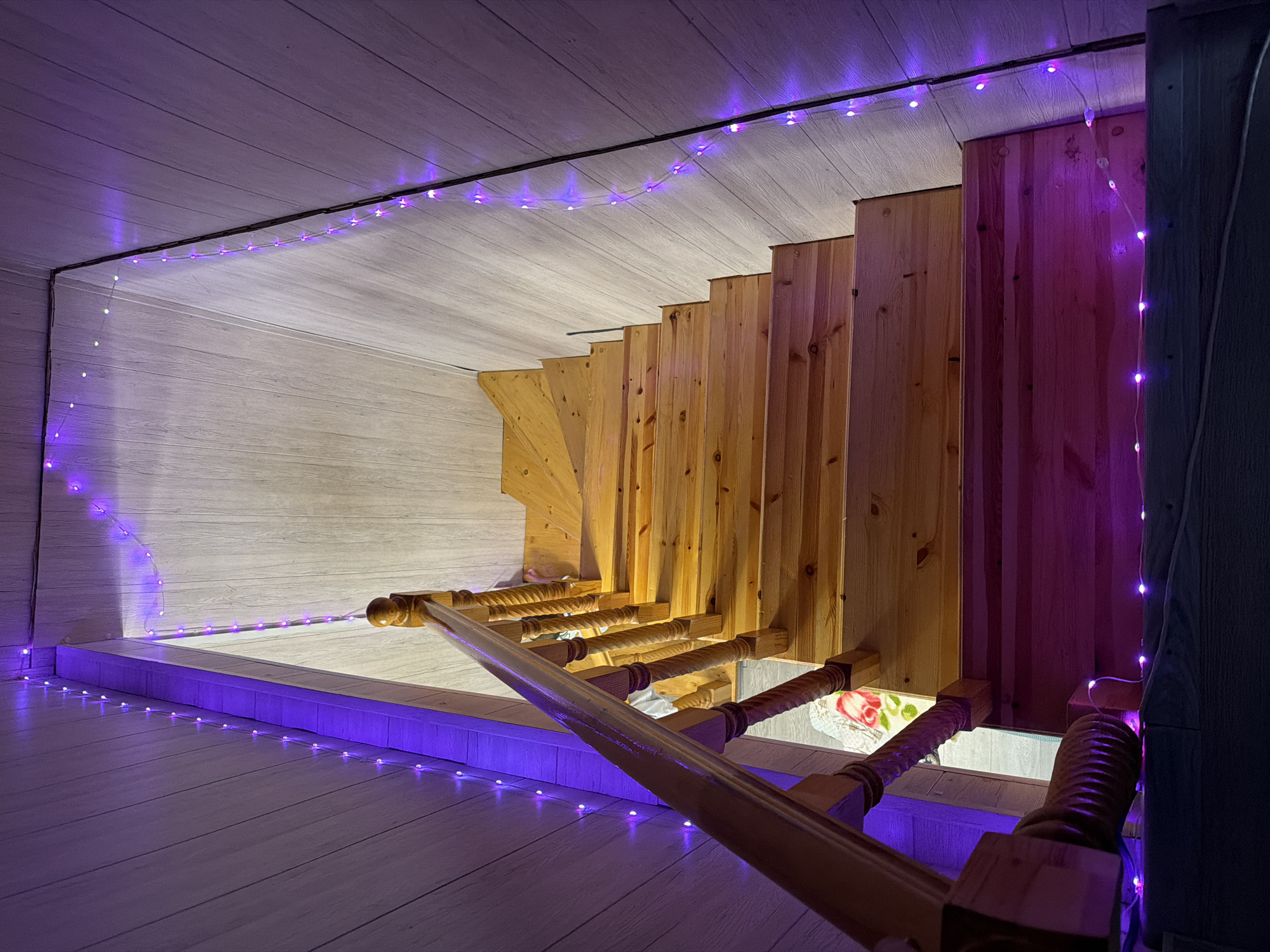 The image shows a wooden staircase with purple string lights running along the walls and the edge of the stairs. The walls are paneled with light-colored wood. A wooden banister with turned spindles is visible on the left side of the stairs. The perspective is from the top of the stairs looking down. The lighting creates a warm and festive atmosphere.