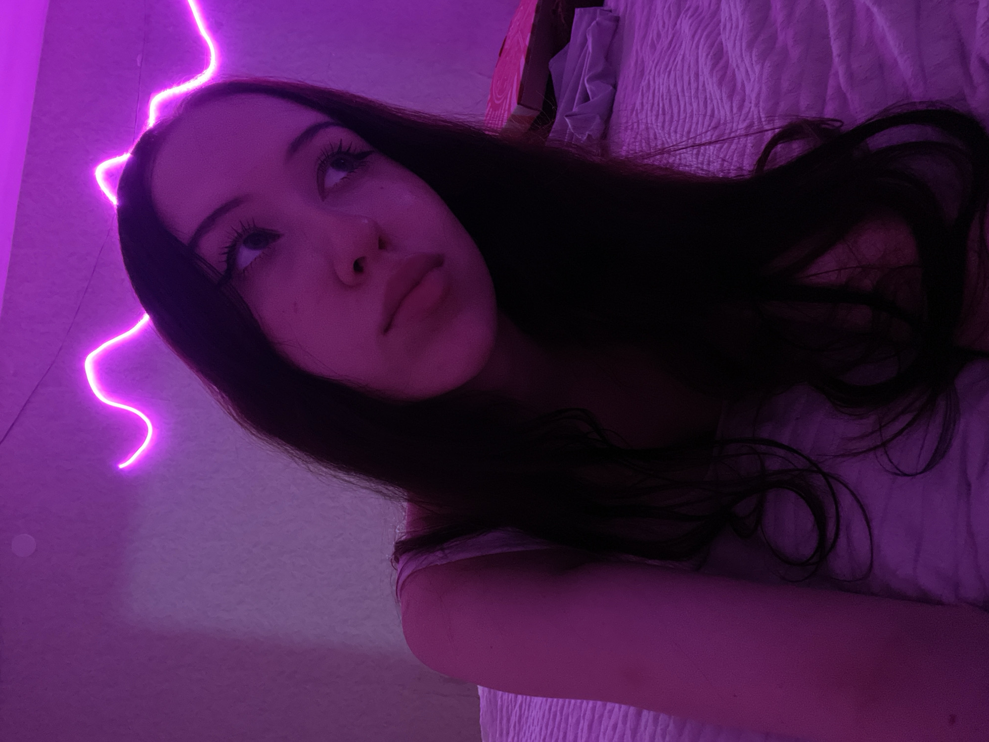 The image shows a young woman with dark hair, looking upwards and to the right. She is wearing a light-colored top. She is lying on a bed with a textured white comforter. Behind her, a pink neon light strip is mounted on the wall, creating a wavy pattern. The room is dimly lit, with the neon light casting a purple glow. To the right of the woman, on the bed, is a pink box and some folded fabric. The scene appears to be indoors, possibly a bedroom. The location is not specified in the image itself.