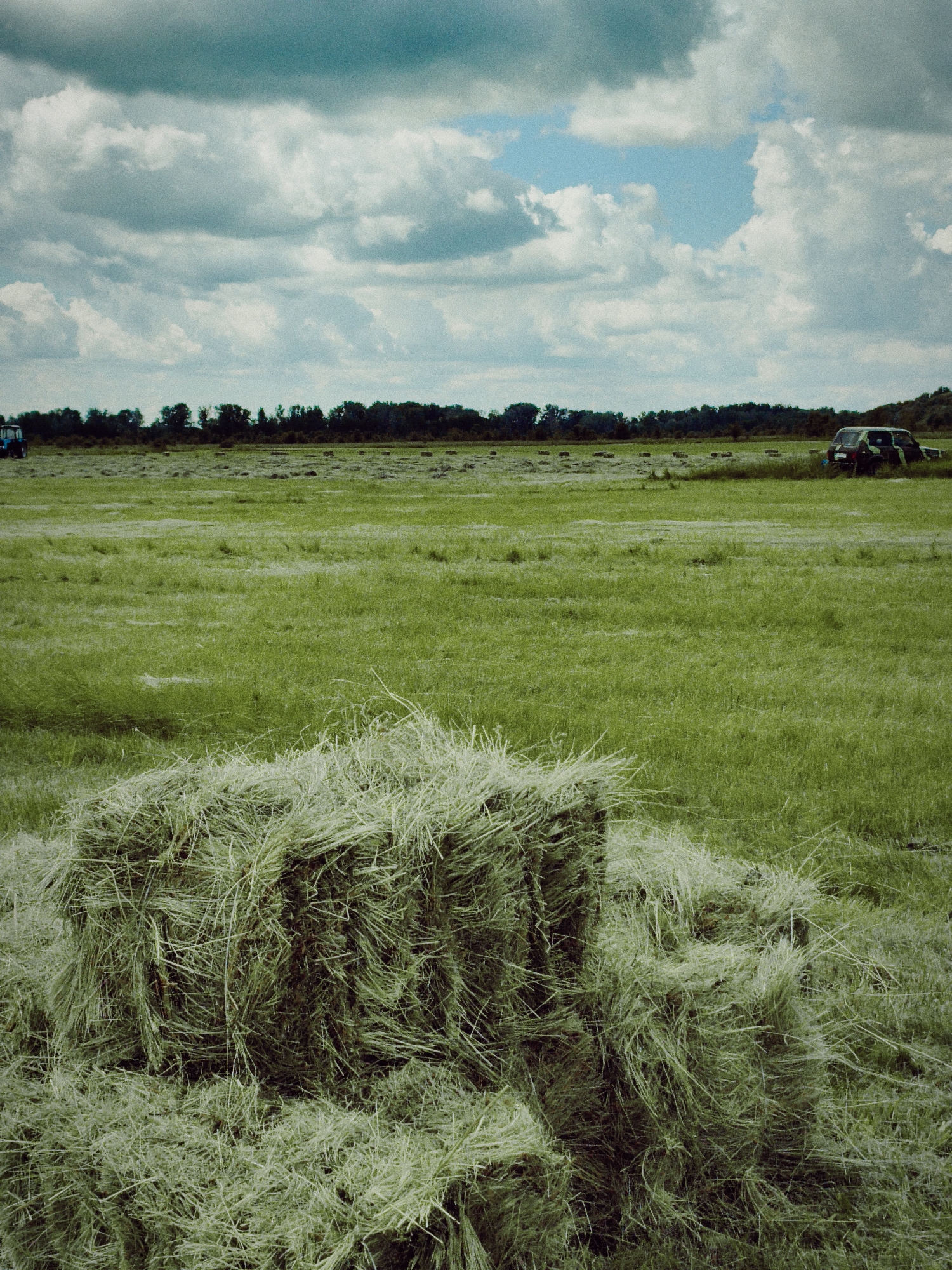 In the foreground of a vast, sunlit green field, a large, rectangular bale of hay rests, partially surrounded by loose, wispy strands of cut grass. The field stretches into the distance, dotted with smaller bales of hay and a dark line of trees bordering the horizon. Above, a dramatic sky unfolds, filled with a mix of dark, textured clouds and patches of bright blue. In the background, a camouflage-patterned vehicle is parked in the field, and a blue tractor is visible on the far left, hinting at agricultural activity. The overall scene depicts a rural landscape in the process of harvest.
