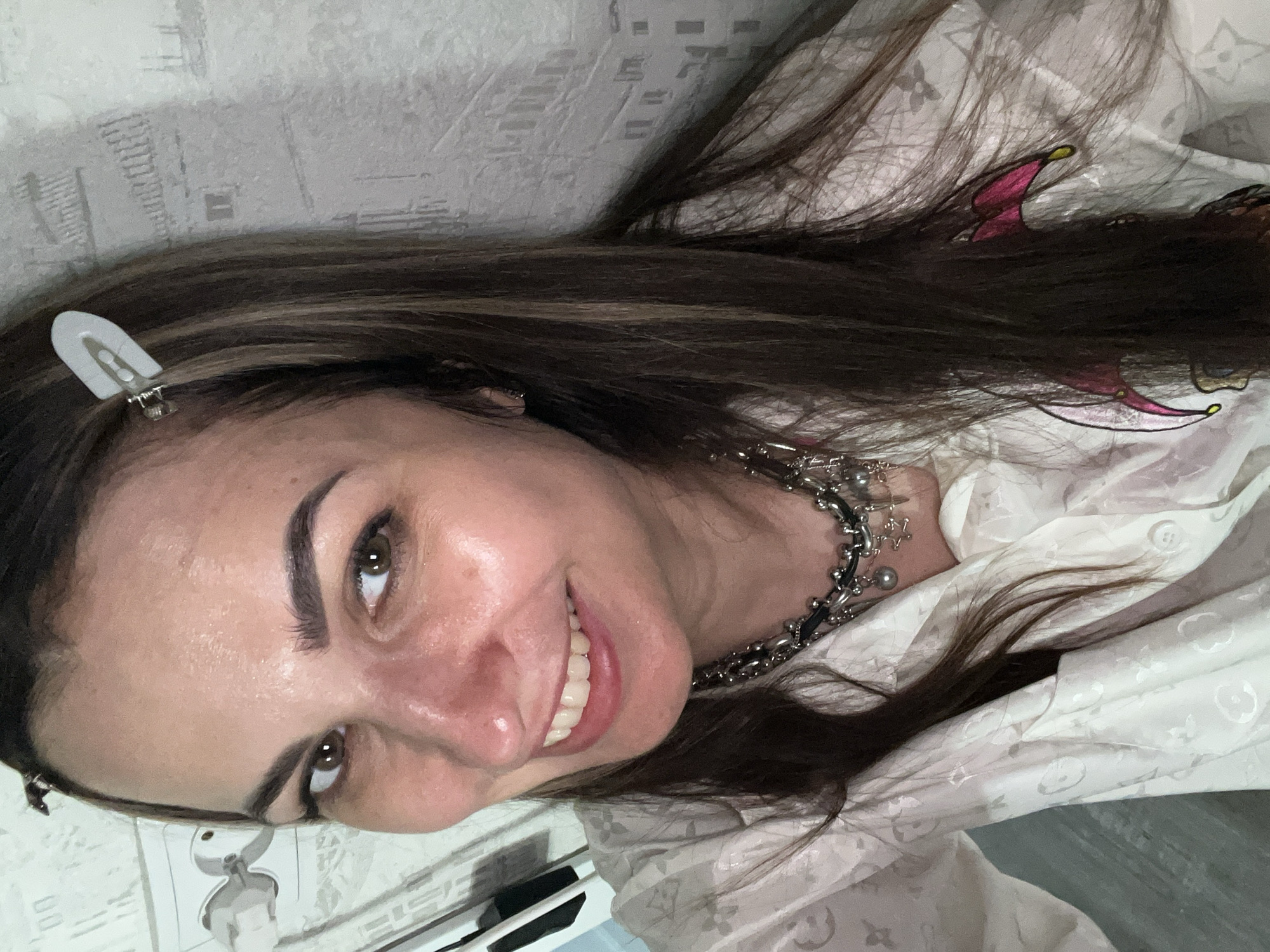 A woman with brown hair smiles at the camera. She is wearing a silver necklace and a white patterned shirt. There is a white hair clip in her hair. The background consists of a patterned wall and what appears to be a white desk or counter with items on it. The location is Unknown, Unknown.