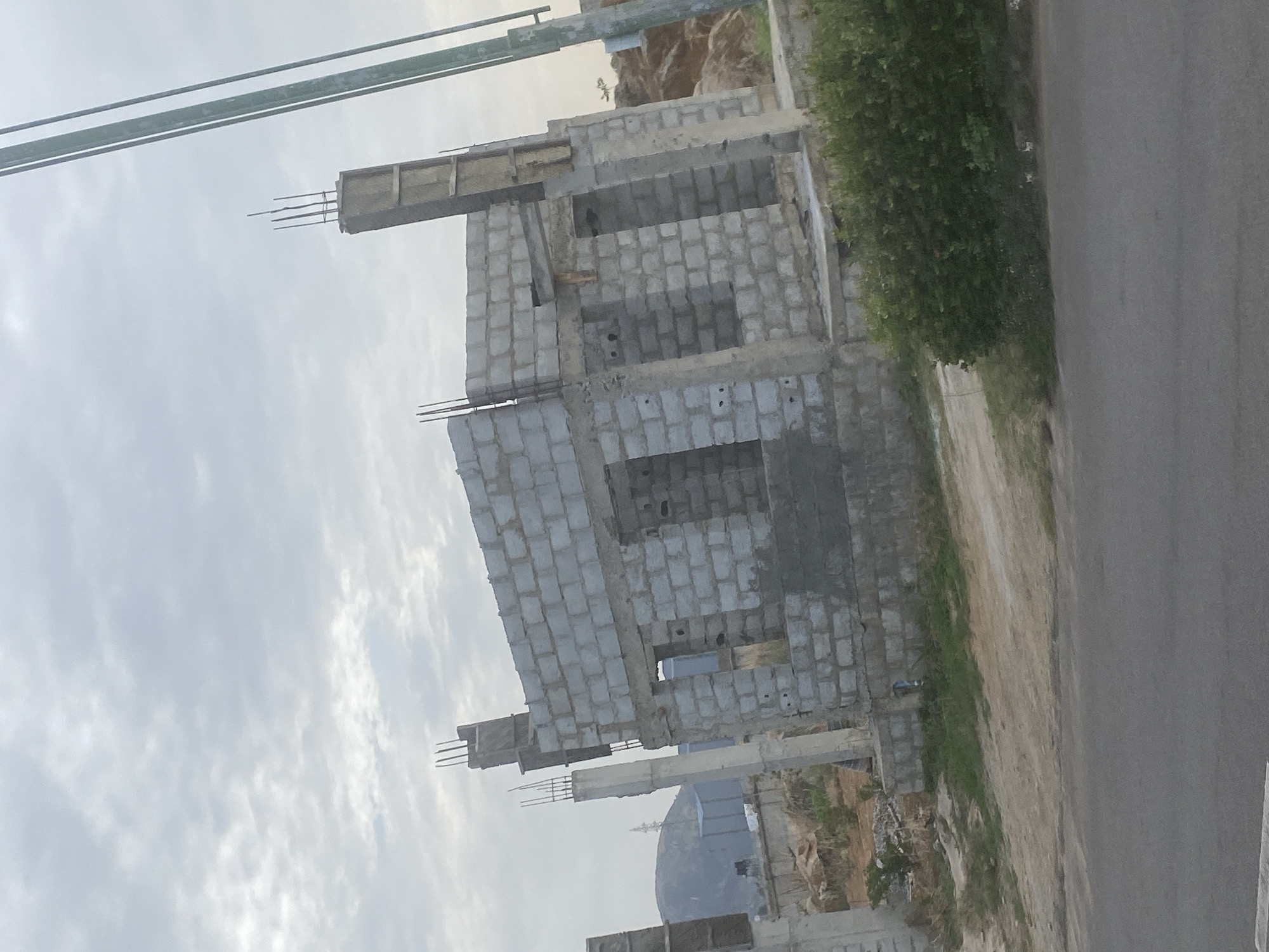 The image shows a building under construction in Abuja, Nigeria. The building is made of grey concrete blocks and has multiple stories. Steel reinforcing bars protrude from the top and sides of the structure. The surrounding environment includes a road and vegetation. The sky is overcast.
