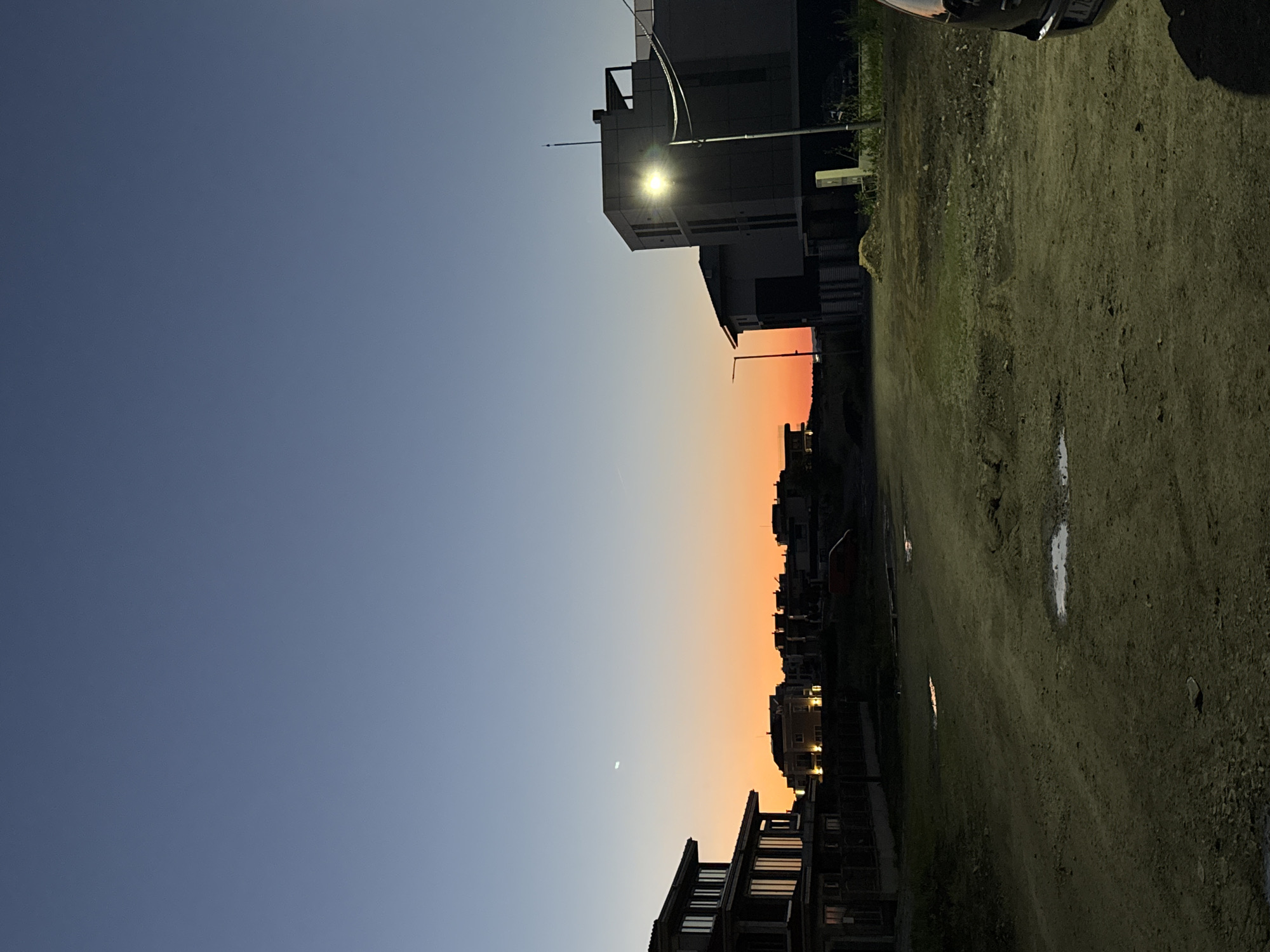 The image depicts a street scene in Burgas, Bulgaria, at dusk or dawn. The sky transitions from a deep blue at the top left to an orange hue on the horizon where the sun is setting or rising. Buildings line the horizon, silhouetted against the orange sky. A crescent moon is visible in the sky. On the right side of the frame, a dirt road or field stretches into the distance. A street light illuminates a building on the right side. Puddles are scattered across the road. A car is partially visible in the top right corner.