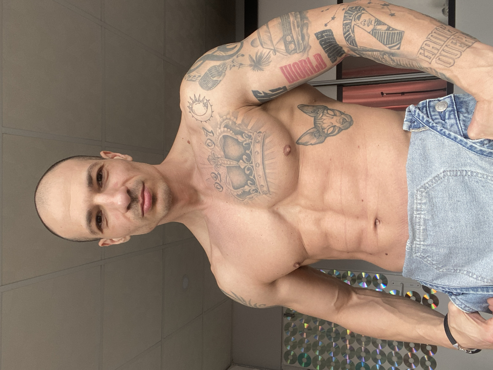The image depicts a shirtless male individual standing in an indoor setting. The subject, who has a shaved head, light complexion, and a muscular physique with visible abdominal definition, looks directly at the camera with a neutral expression. His chest, abdomen, and arms are covered with numerous tattoos. On his upper chest, a large crown design with radiating lines is visible, along with the numbers "02" and "07." A Sphynx cat head is tattooed on his left ribcage. His right arm displays various tattoos, including a sun and crescent moon, a microphone, abstract shapes, a classic car, a palm tree, text blocks (one appearing to read "MONEY"), an Egyptian pharaoh's head within the number "7," stars, and the phrase "GAME OVER." The individual wears light blue denim overalls, unfastened and pulled down to his mid-waist, exposing his torso. His right hand is inserted into the waistband or pocket of the denim garment. A black wristband is present on his right wrist.
The background features a light-colored, gridded drop ceiling with one partially visible recessed light fixture. To the subject's left, a wall or panel is covered with a uniform arrangement of reflective, circular objects, likely compact discs. To his right, a dark, reflective surface, consistent with a mirror or closet door, is visible, with a faint reddish reflection or internal color behind it.
The image depicts an indoor scene; however, specific visual cues identifying the location as Pleven, Bulgaria, are not present within the frame.