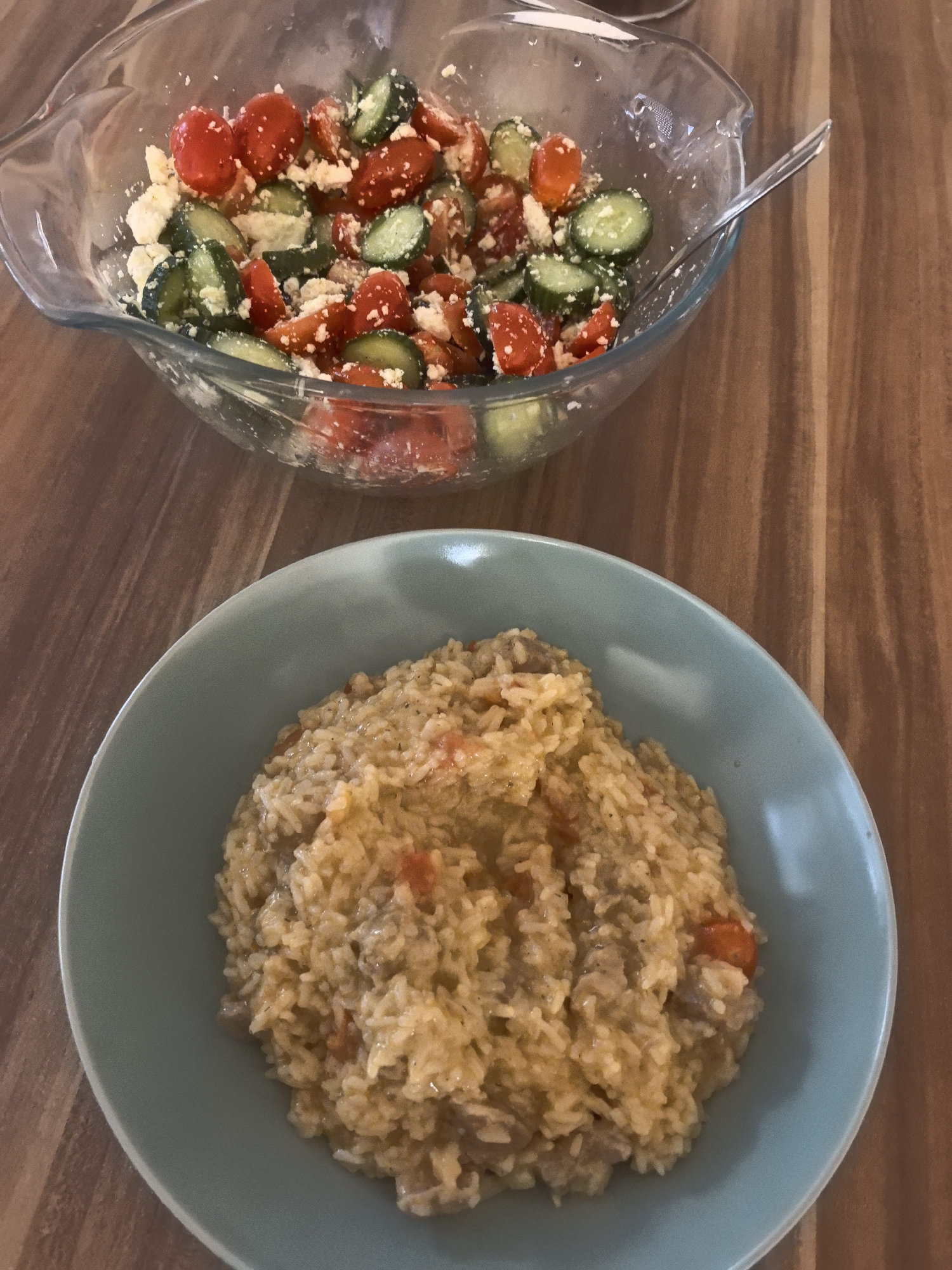 The image displays a home-cooked meal arranged on a wooden table, likely a dining table or kitchen counter in Hattersheim am Main, Germany. In the upper part of the frame, a clear glass bowl is filled with a fresh salad. The salad consists of halved red cherry tomatoes, sliced green cucumbers, and crumbled white cheese, possibly feta. A clear serving spoon is partially visible within the salad. Below and in the foreground, a round, light blue-green ceramic bowl holds a savory-looking rice dish, resembling risotto or a rice pilaf. This dish has a creamy, yellowish-brown texture, interspersed with small red pieces, likely tomatoes, and possibly other ingredients such as meat or vegetables. The wooden surface beneath the bowls exhibits a distinct grain pattern. The scene depicts a meal prepared for consumption, without any people present. No text is visible in the image, and the time of day is not explicitly indicated.