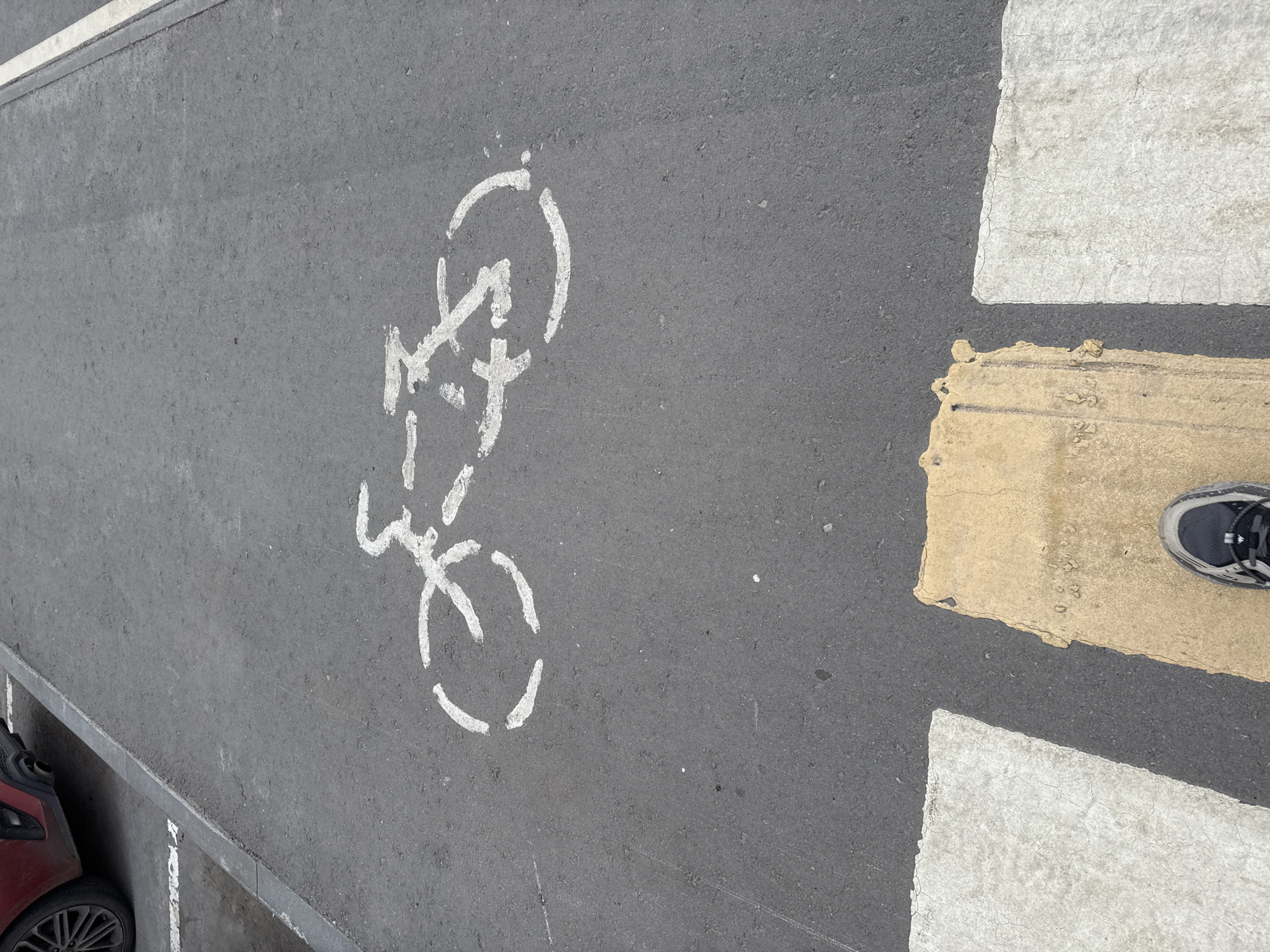 An aerial view of a paved road shows a white bicycle stencil painted on the asphalt. The stencil depicts a simplified bicycle with two wheels and a frame. In the foreground, a portion of a crosswalk is visible, with alternating white and yellow painted sections. A person's foot, clad in a dark-colored athletic shoe, is positioned on the yellow section of the crosswalk. To the left of the image, the rear of a dark red car is partially visible, with its wheel and part of the bumper showing. The environment appears to be an urban setting.