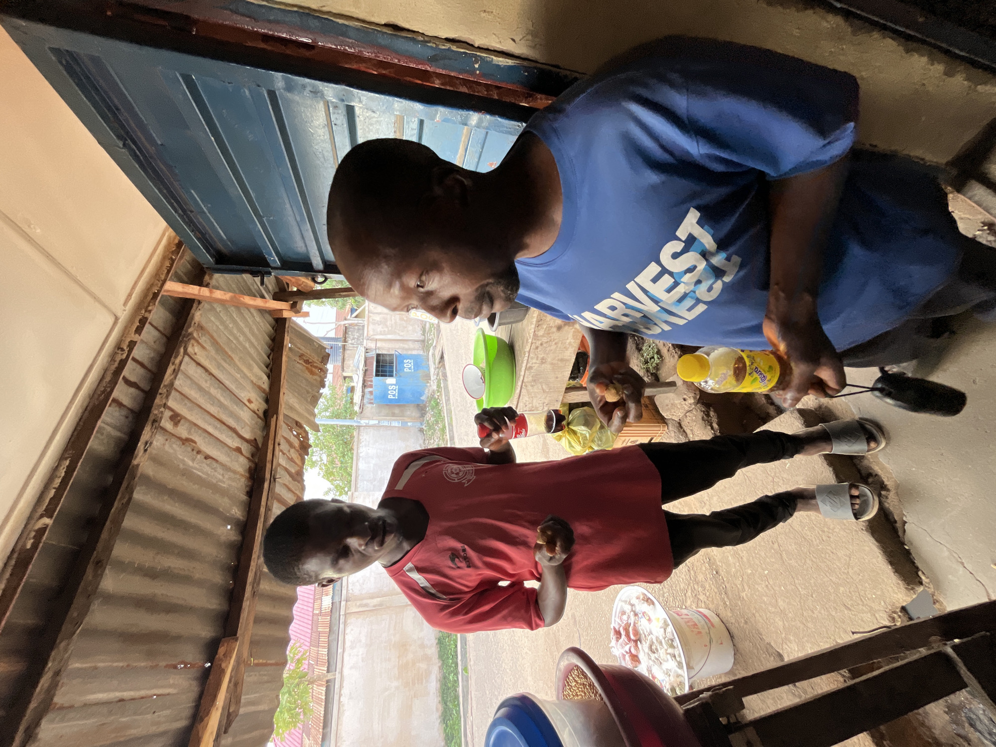 Two men are preparing food in an outdoor setting. The man on the right wears a blue shirt with "HARVEST" printed on it. He's holding food items. The man on the left wears a red shirt and holds food as well. There are containers with ingredients around them. The location appears to be a roadside or open-air cooking area, possibly in Jos, Nigeria, as indicated by the surroundings and possible building styles. The environment includes corrugated metal structures and basic food preparation items.