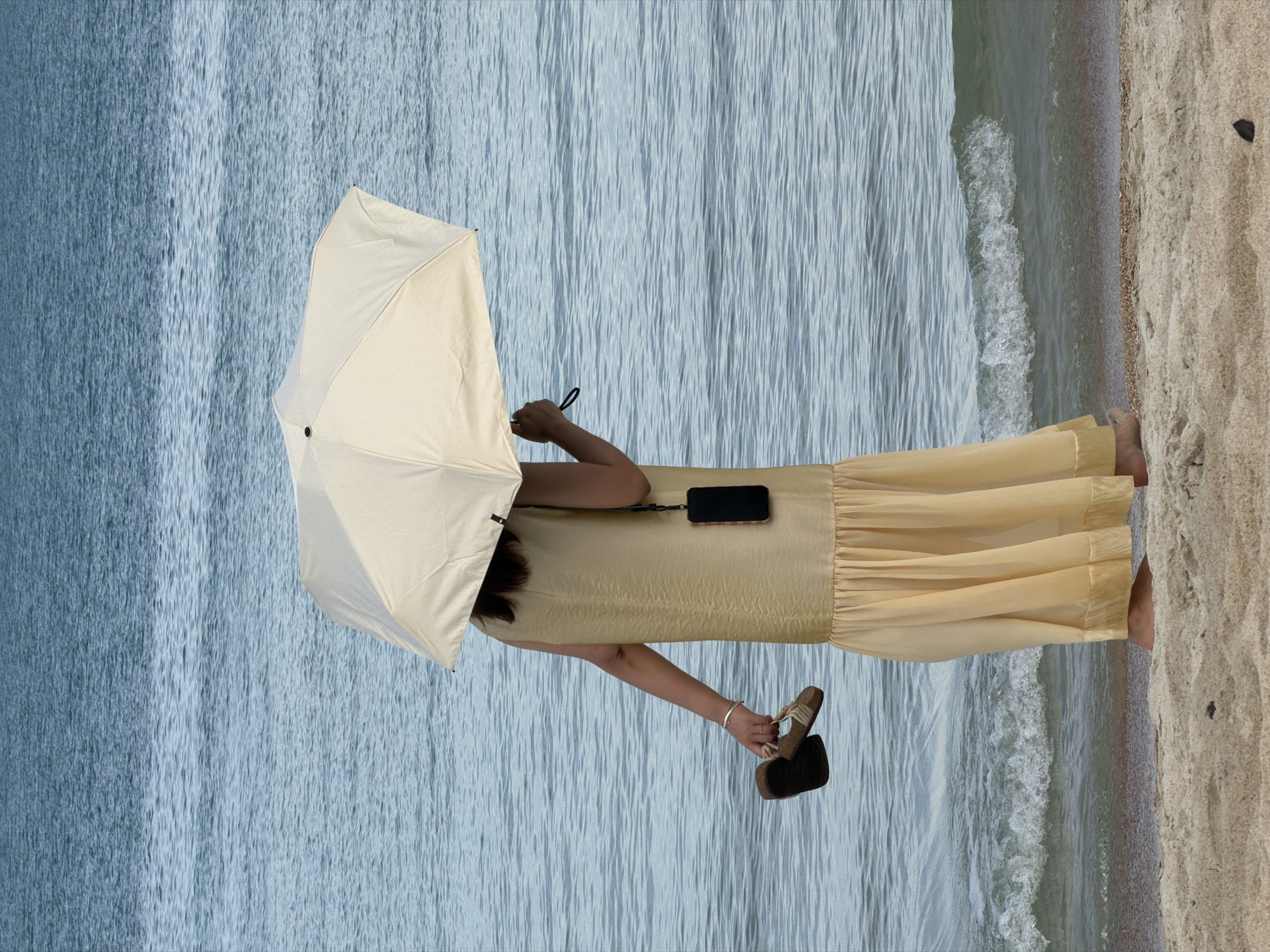A person stands on a sandy beach in front of a calm sea, holding a light-colored umbrella over their head. The person is wearing a long, flowing golden dress and is barefoot, with sandals held in their left hand. A smartphone is attached to a lanyard around their neck. The water is a deep blue, with gentle waves lapping at the shore. The sand is light brown and textured. The scene appears to be during daylight hours. The location is identified as Baan Chaweng, Thailand.