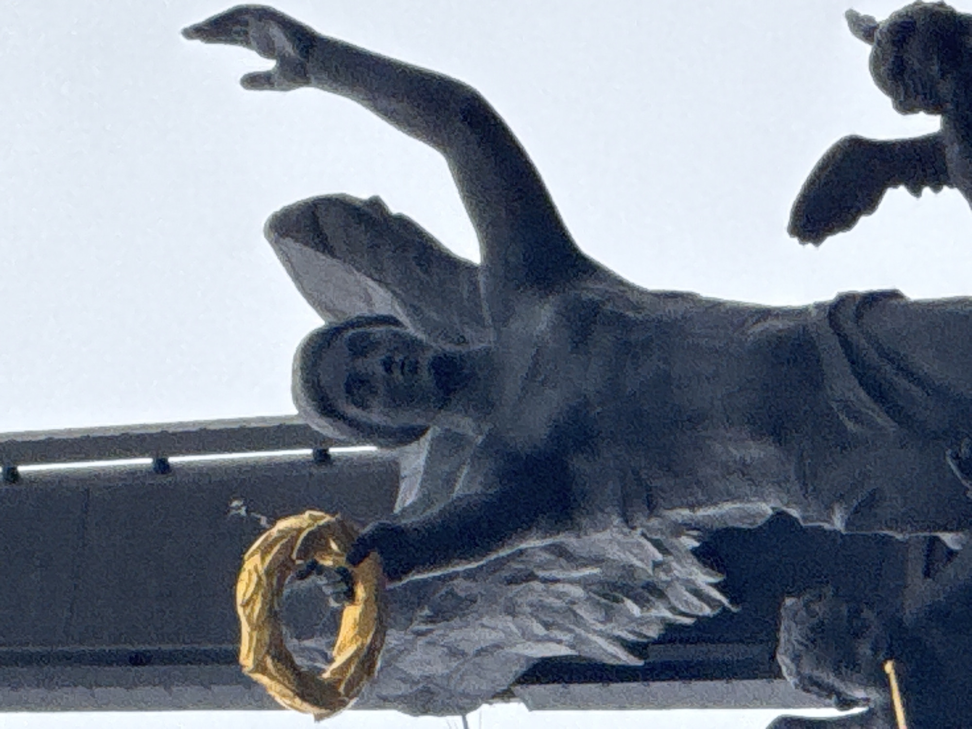 The image shows a close-up of a dark, weathered statue of an winged figure, possibly an angel or victory figure, holding a golden wreath in its left hand. The statue is part of a larger monument, with a dark, vertical structure visible behind it. The figure's right arm is raised, with the hand open and facing upwards. The statue is dark, appearing to be made of bronze or a similar metal, and shows signs of age and weathering. The background is a pale, overcast sky. Parts of other statues or figures are visible at the bottom of the image, including what appears to be a child's head on the left and wing-like appendages on the right, suggesting a larger sculptural group. This monument is likely the Monument to the Conquerors of Space, located in Moscow, Russia.