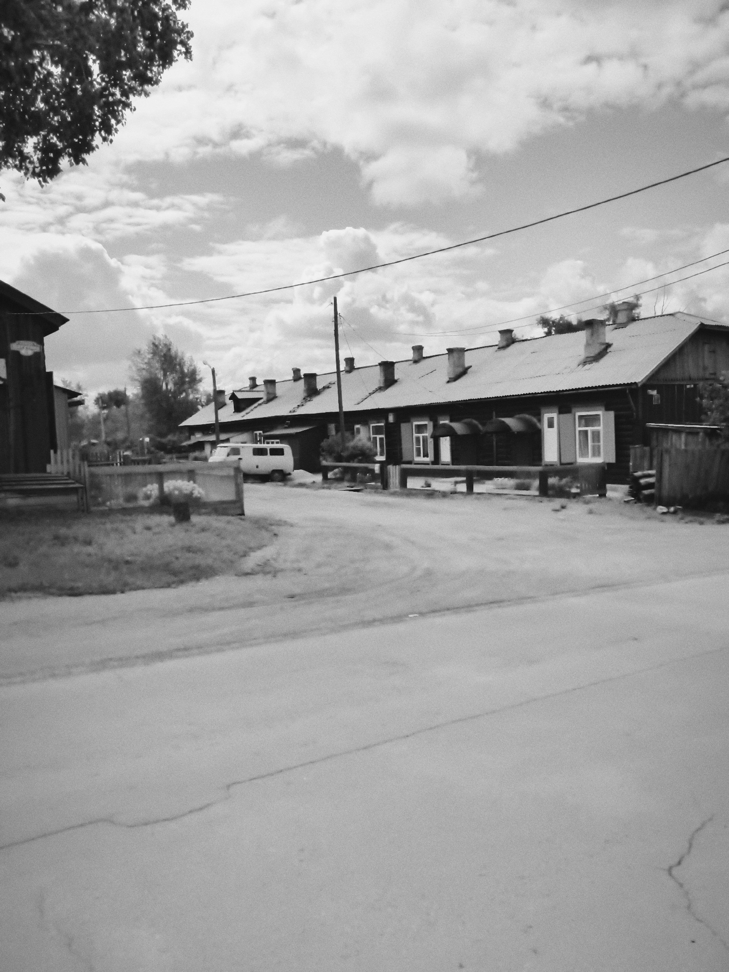 In Cheremkhovo, Russia, a black and white, medium-shot photograph captures a street scene with a row of wooden houses. The houses, characterized by their distinctive peaked roofs and numerous chimneys, are situated along a paved road. In front of one of the houses, a white van is parked. A wooden fence borders a grassy area in the foreground. The sky above is filled with scattered clouds. The overall atmosphere of the image is serene and rustic.