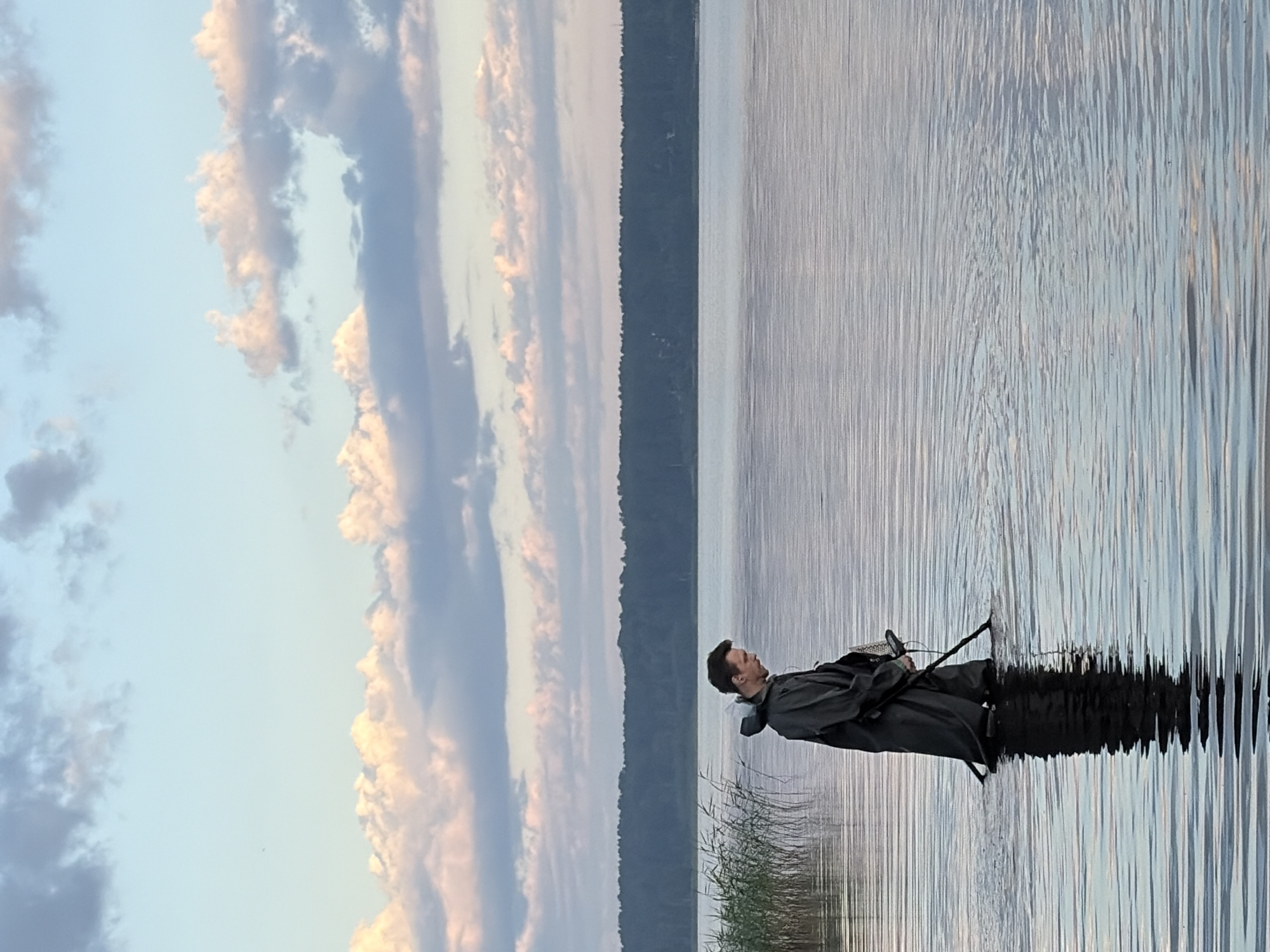 A person is metal detecting in shallow water on a large lake, with a treeline visible in the background. The sky above is a pale blue with scattered pink and white clouds. The water is calm, with ripples reflecting the sky. The person is wearing waders and a dark jacket, and is holding a metal detector. The image appears to be taken at dusk or dawn due to the lighting.