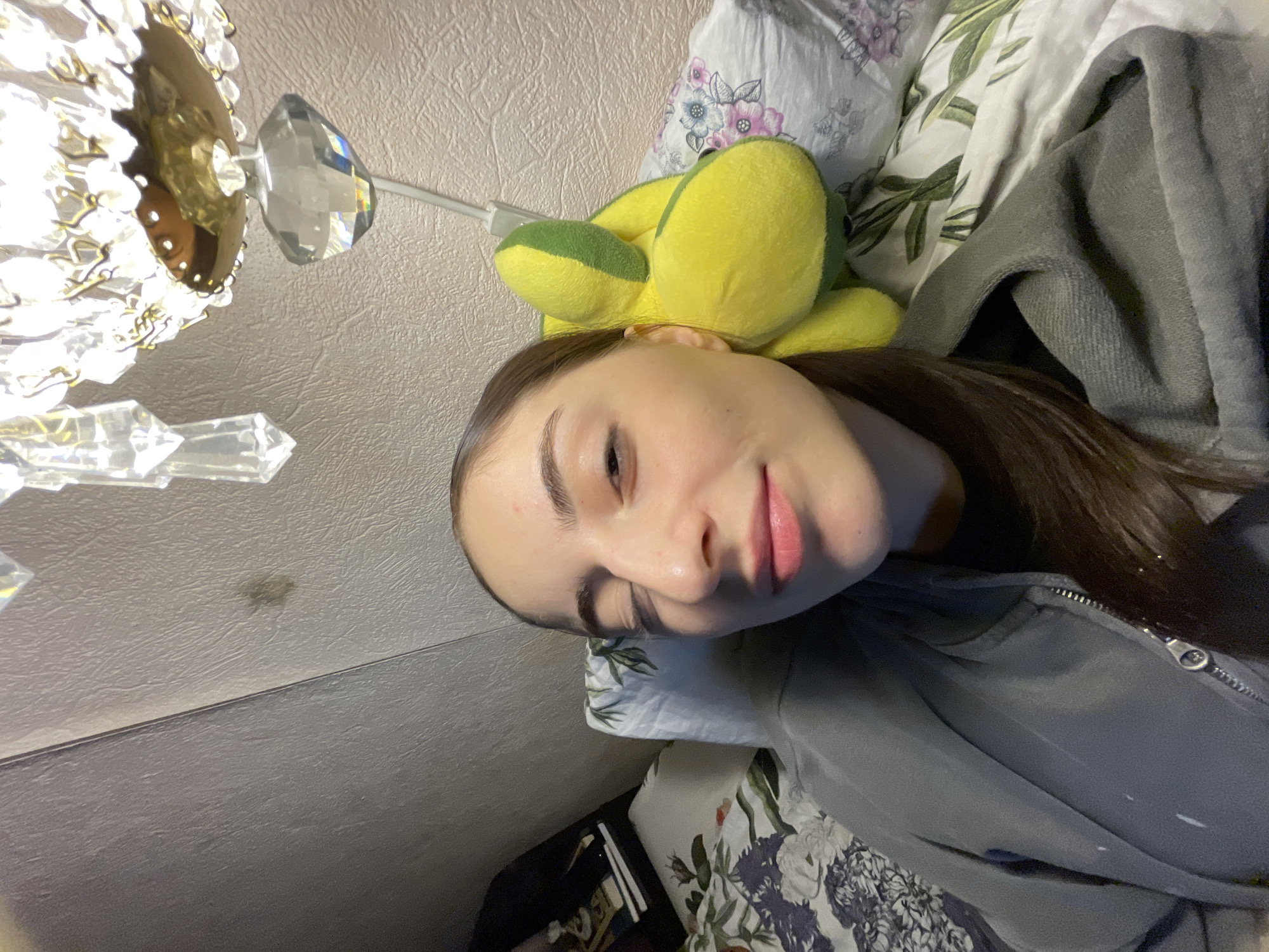 The image displays a close-up, slightly high-angle view of a female individual lying down in an indoor residential setting in Moscow, Russia. The individual, with fair skin and dark hair, has her eyes partially closed and a subtle smile. She is dressed in a gray zippered hoodie over a dark garment. Her head rests on a pillow featuring a floral pattern of blue and purple flowers on a white background. Behind her, on a second pillow with a green leaf pattern, is a yellow and green plush toy. In the upper right quadrant, a portion of an illuminated crystal chandelier with gold-toned metal accents is visible. The background consists of light-colored, textured wallpaper, which exhibits a small, dark, irregular smudge above the individual's head. A dark furniture item with stacked objects is partially discernible on the left side of the frame.