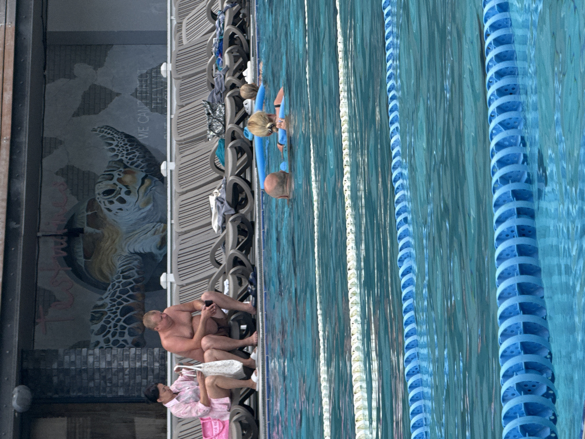 In Yalta, Ukraine, a scene unfolds at an outdoor swimming pool complex. A woman in a pink patterned shirt and a man in swimming trunks sit on lounge chairs near the pool's edge. The woman holds a white bag and a mobile device, while the man, with a phone in his hand, appears to be looking at it. In the background, a mural of a sea turtle adorns a wall, with the words "WE CAN'T DIRECT THE WIND BUT WE CAN ADJUST OUR SAILS" partially visible. Further into the pool, two individuals are swimming, one using a blue pool noodle. The pool is divided by lane markers, with blue lane dividers visible in the foreground. The overall environment suggests a recreational setting on a clear day.