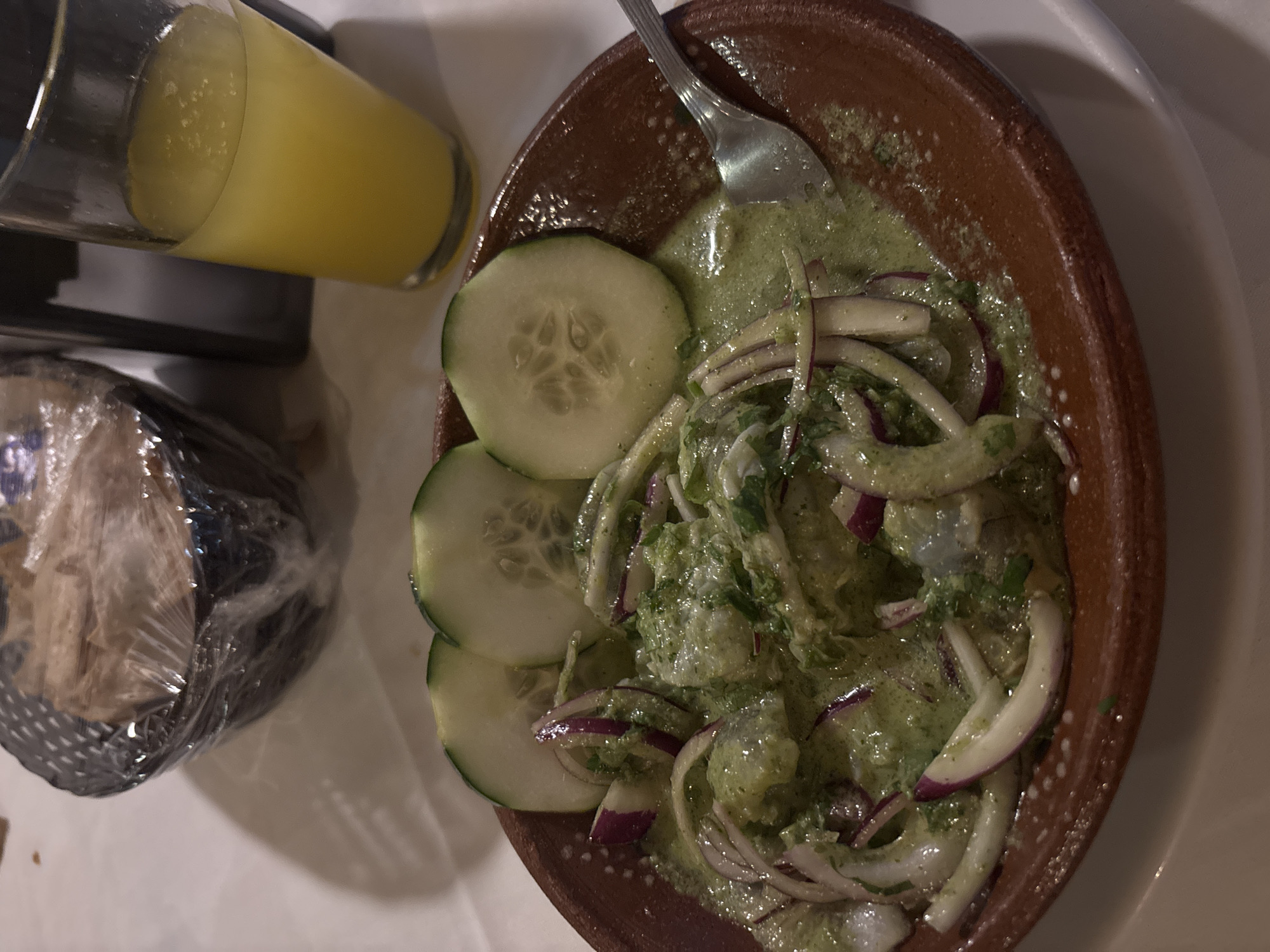 A still-life photograph shows a plate of ceviche in a terracotta-colored bowl alongside a glass of yellow liquid and a wrapped bundle, likely tortillas or bread, set on a white surface.
The ceviche consists of what appear to be shrimp or white fish mixed in a green sauce, garnished with sliced red onions and cucumber rounds. A fork rests in the ceviche. The terracotta bowl has a simple, rustic appearance.
The glass contains a yellow beverage, possibly juice or a mixed drink, and sits next to a dark object, perhaps a coaster or tray. The wrapped bundle is partially visible, encased in clear plastic. The white surface is likely a table. The overall composition suggests a casual meal setting, possibly in Veracruz, Mexico, given the prevalence of ceviche in the region. No people are visible in the frame.