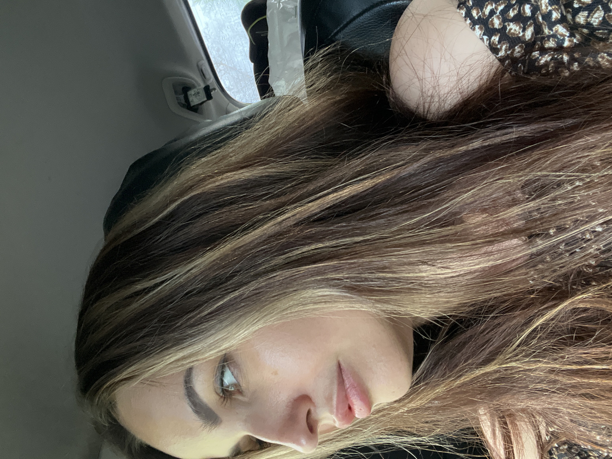 A woman with long, brown hair with blonde highlights is in the passenger seat of a car. She is looking out the window to her left. The interior of the car is visible, including the headrest of the seat in front of her and the ceiling of the car. A small black object is hanging from the ceiling, possibly a dashcam or rearview mirror. The background outside the window is blurry, suggesting movement. She is wearing a black top with a leopard print trim on the shoulder.