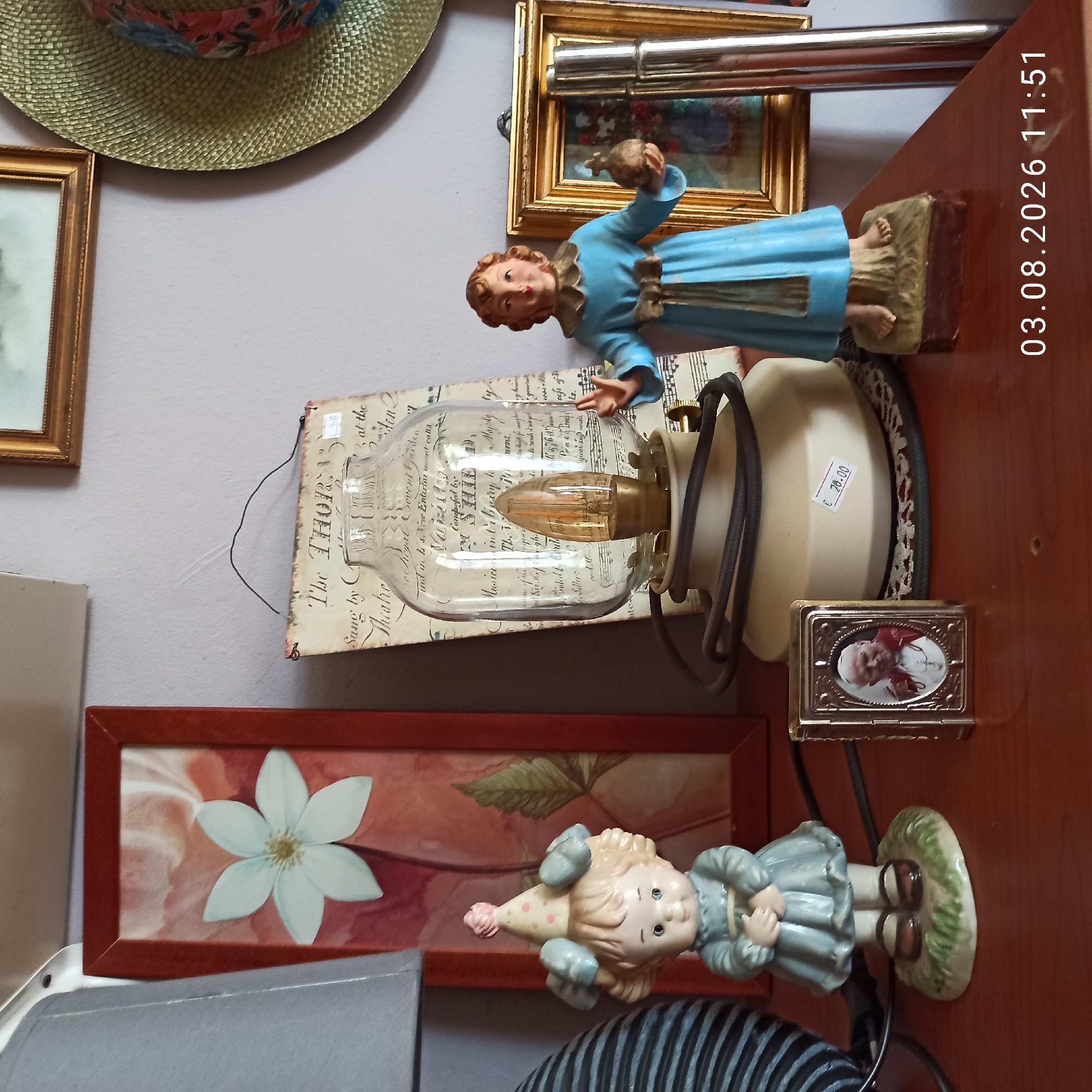 The image depicts a collection of decorative items displayed on a wooden surface. On the left, a ceramic figurine of a young girl wearing a blue dress and a party hat stands on a patch of green grass. To her right is a small, ornate golden frame containing a miniature portrait of Pope John Paul II. Behind these items, a vintage oil lamp with a glass shade and an exposed filament bulb is prominent. The lamp sits on a crocheted doily, and a price tag displaying "€ 28.00" is visible on its base. Further to the right, a larger figurine of a child in a blue robe stands on a rectangular base, holding a small sphere. Behind this figurine, a vintage picture frame with a golden ornate border encloses a painting of a floral arrangement. A woven straw hat with a colorful fabric band rests on the wall in the upper right corner. A framed painting of a white flower in a reddish-brown frame is positioned on the left side of the image, behind the girl figurine. The wall appears to be a neutral, light color. Text is present on a piece of aged paper with musical notes, which is hanging behind the lamp. A date stamp "03.08.2026 11:51" is visible in the lower right corner of the image. The overall scene suggests a curated display of decorative objects, possibly in a home or a shop.
